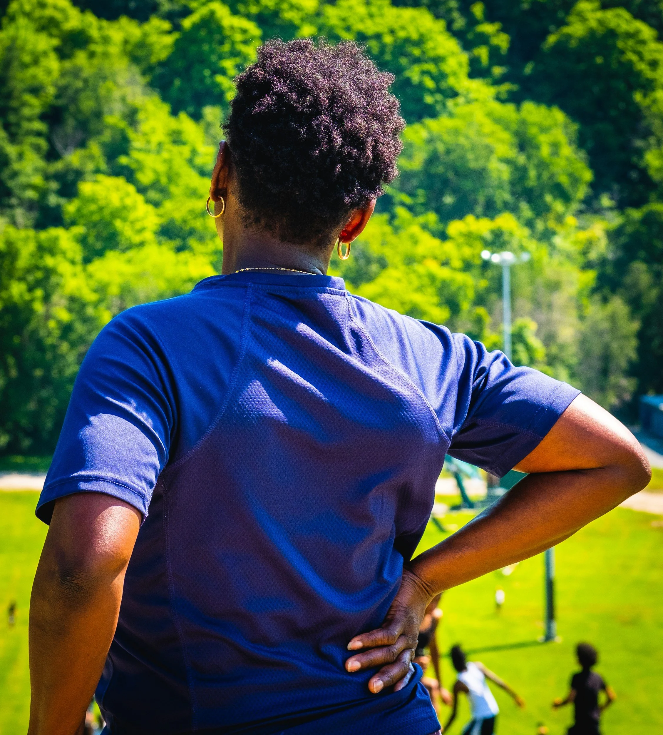 A woman with short curly hair, wearing gold hoop earrings and a blue athletic shirt, stands outdoors with her back to the camera, observing children playing in a grassy park with trees and a soccer goal in the background.