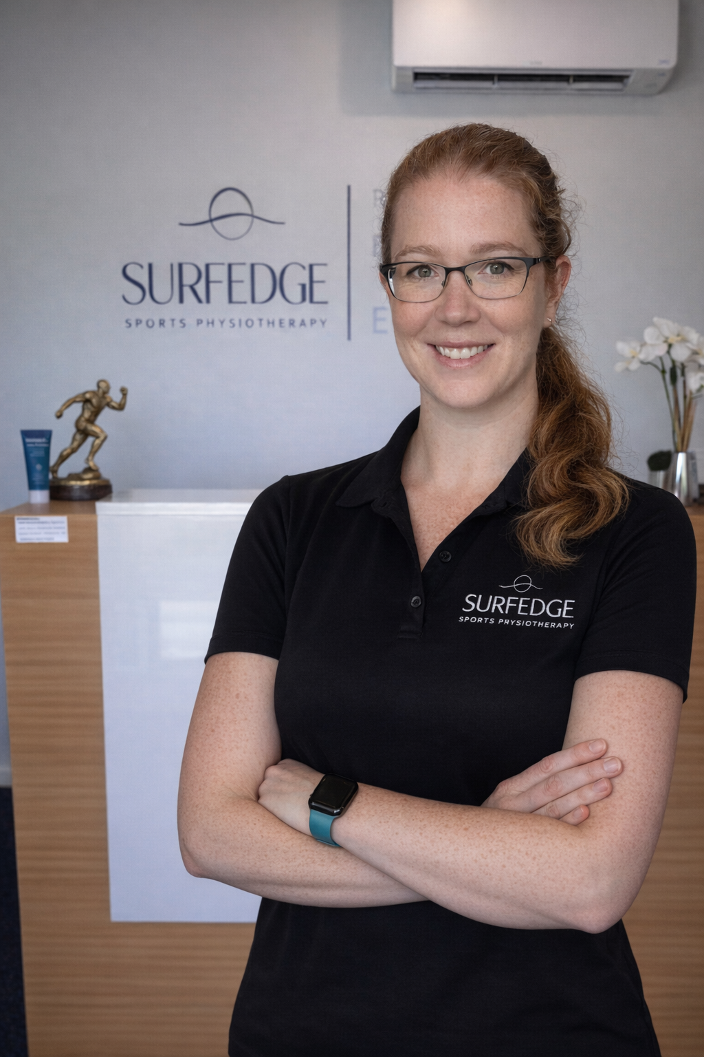 A woman with red hair and glasses smiling at the camera, wearing a black polo shirt with the logo 'SURFEDGE SPORTS PHYSIOTHERAPY,' standing in a reception area with a logo and sports-themed decor.
