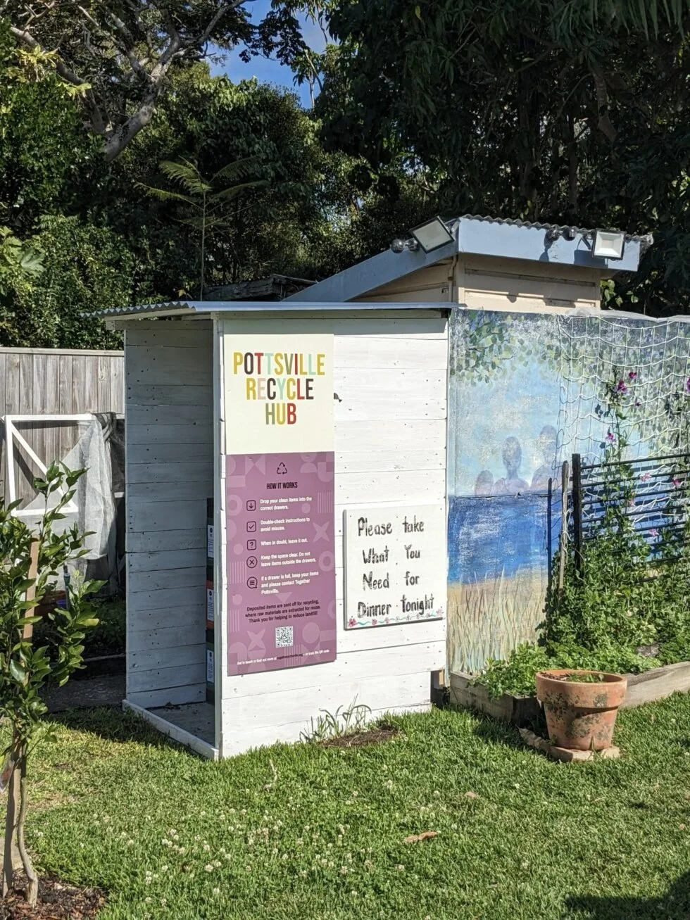 A small outdoor recycling and waste bin station labeled 'POTTSVILLE RECYCLE HUB' in colorful letters, with instructions on how to recycle and a note to take what is needed for dinner tonight, set in a garden with a mural and potted plants.