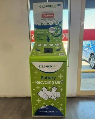 Green EcoBAT recycling station for batteries located inside a store, near a glass door with a parking lot visible outside.