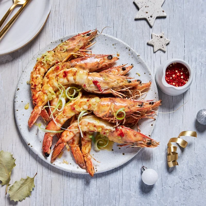 Plate of cooked shrimp garnished with green onions and red pepper flakes, with a side of dipping sauce, set on a white wooden table with Christmas ornaments and leaves around.