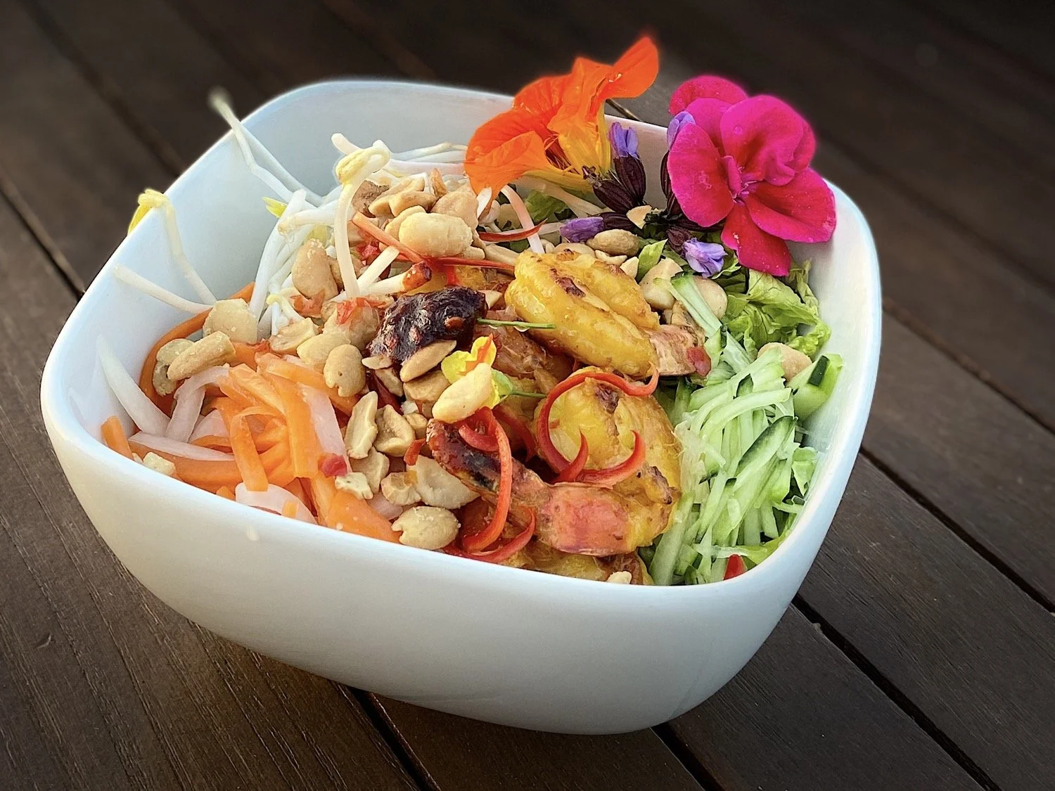 A white bowl filled with a salad containing shredded carrots, bean sprouts, grilled plantains, peanuts, cucumber slices, cherry tomatoes, mixed greens, and edible flowers on top, placed on a wooden surface.
