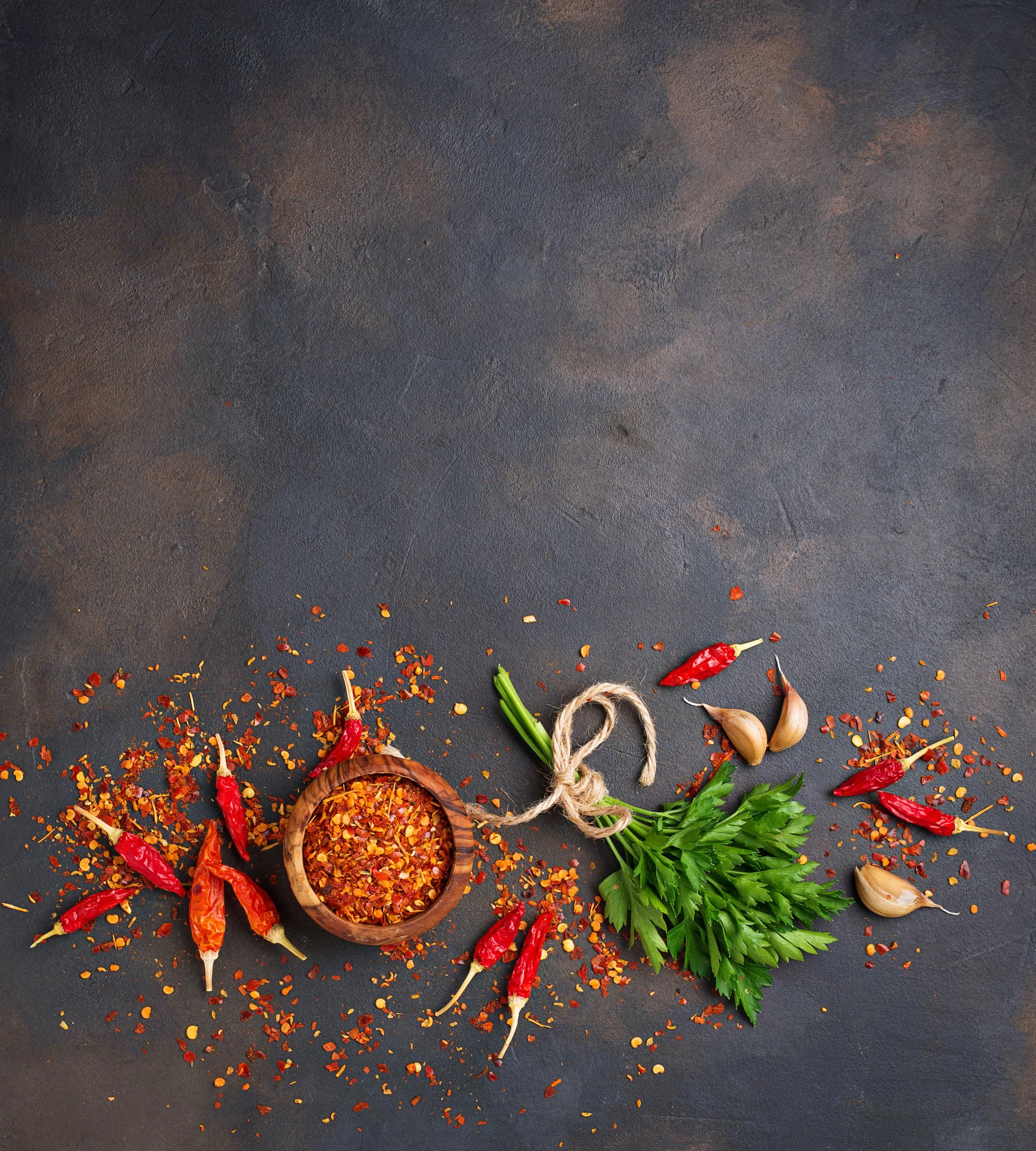 Dried red chili peppers, garlic cloves, fresh parsley, and crushed chili flakes in a wooden bowl on a dark surface.
