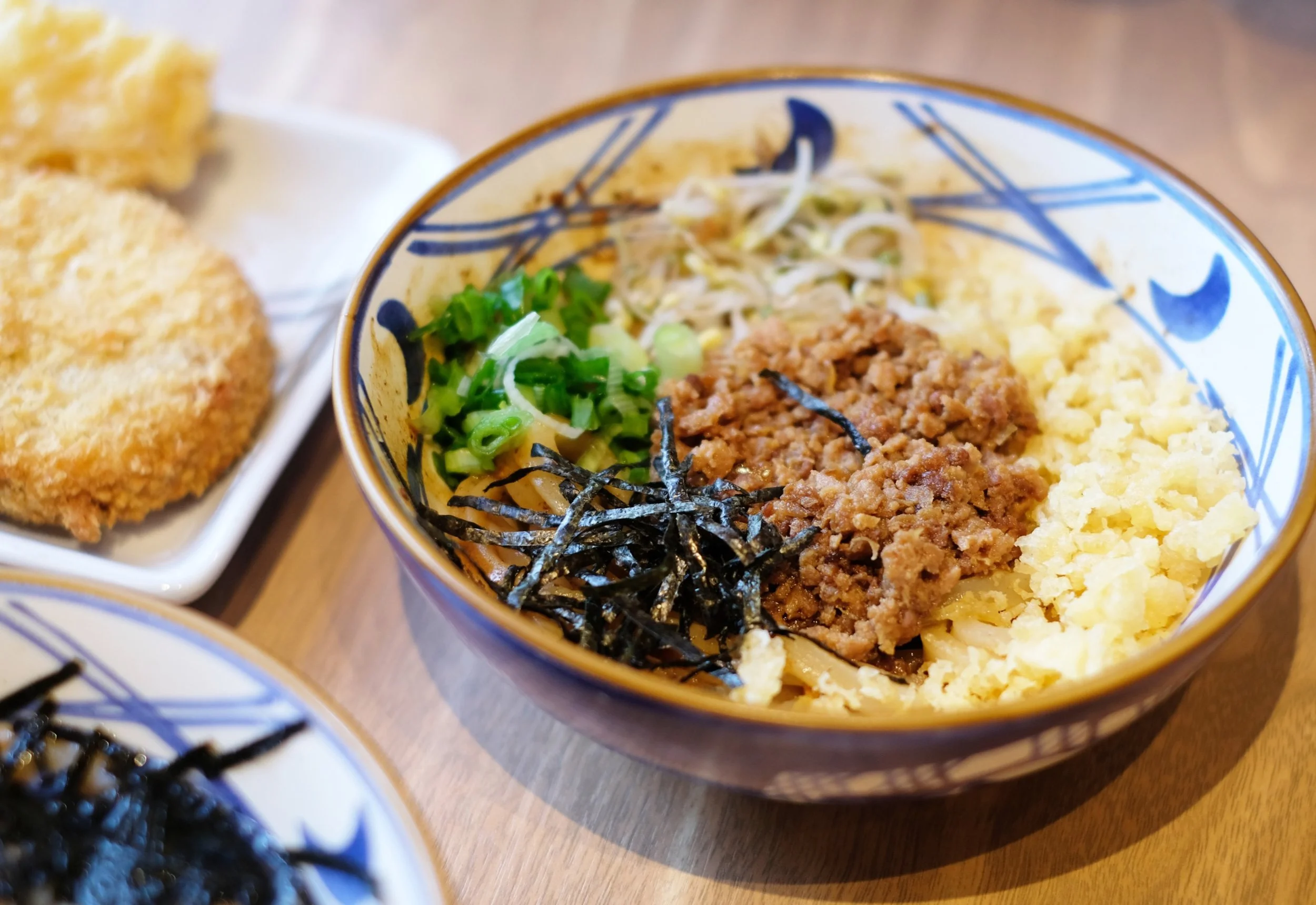 Bowl of Japanese-style rice topped with ground meat, scrambled eggs, chopped green onions, shredded nori, and bean sprouts, with fried cutlets and another bowl visible in the background.