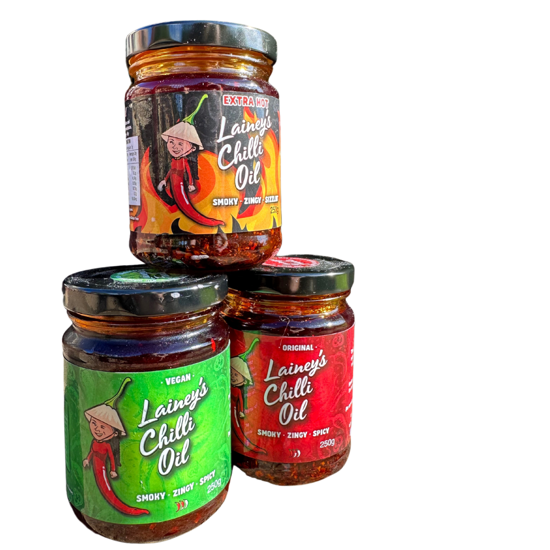 Three jars of Lainey's Chilli Oil, with one labeled extra hot on top and two labeled original and vegan on the bottom, each containing spicy chili oil.