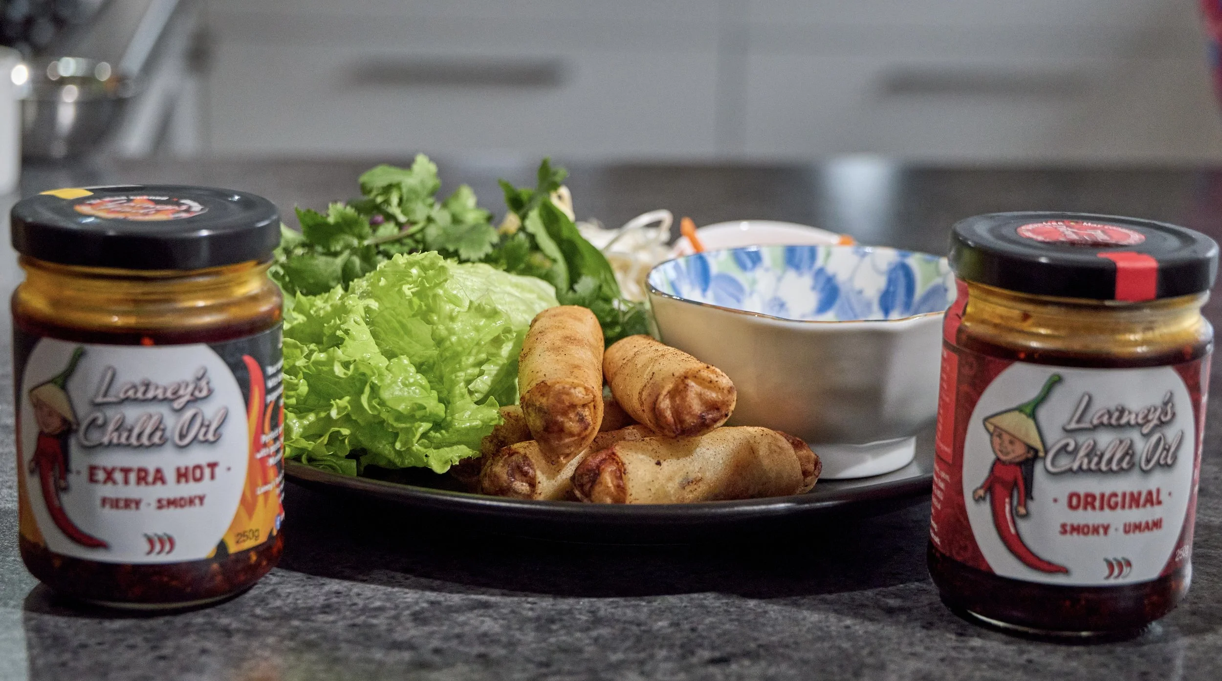 A black plate with fresh green lettuce, cilantro, and two rolled spring rolls, alongside a white bowl with a blue pattern, and two jars of Laurens Chilli Oil, Extra Hot and Original, on a kitchen countertop.