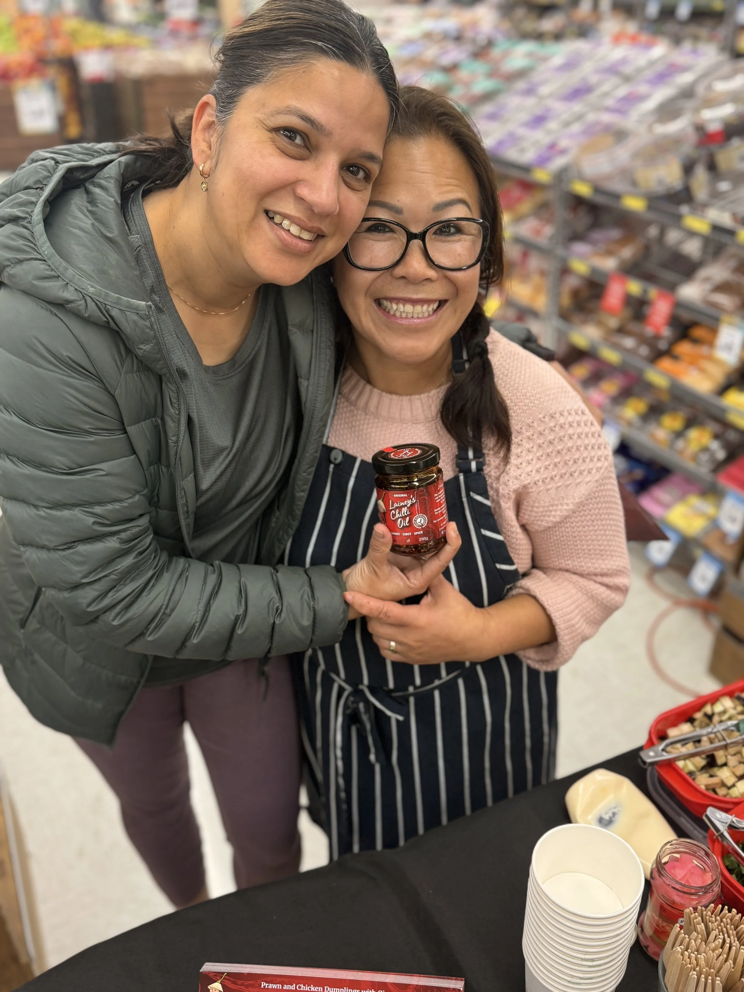 Two women smiling at a grocery store, holding a jar of chili oil with a display of food items and condiments on a table in front of them.