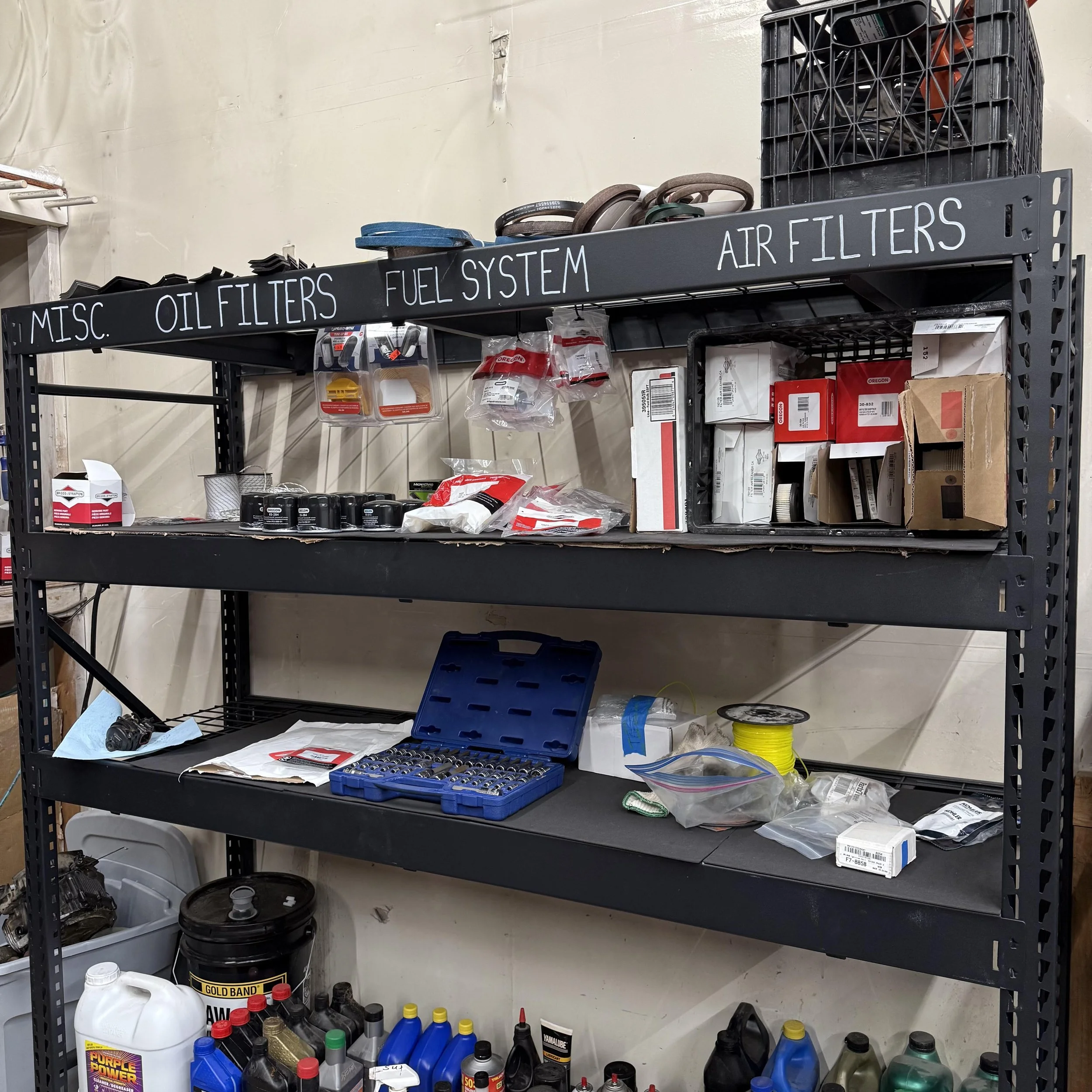 Black metal shelf with labeled sections for miscellaneous items, oil filters, fuel system, and air filters, containing various automotive parts, filters, boxes, and tools.