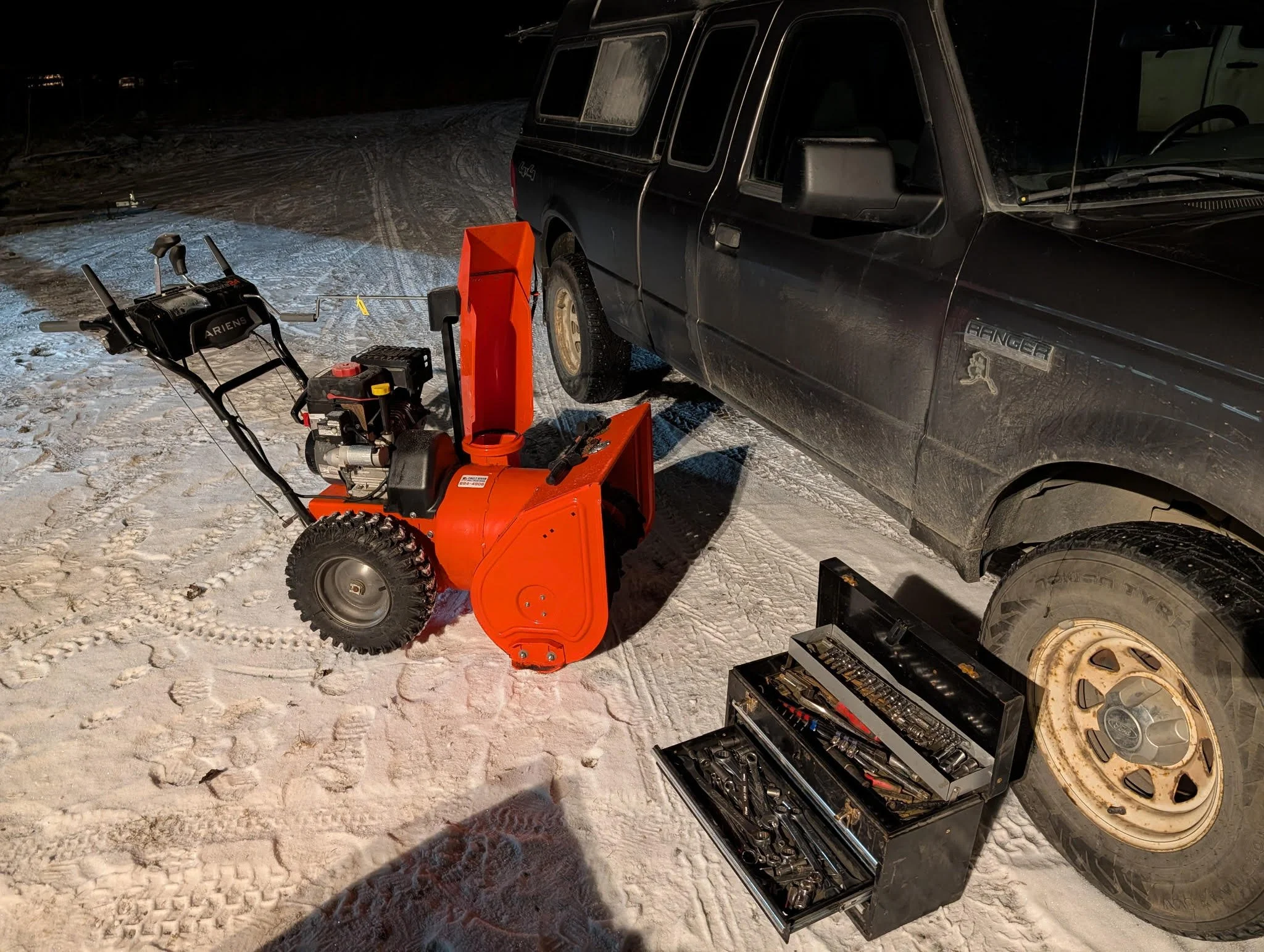 A snowblower within a tool kit and an all-terrain vehicle parked on snow-covered ground at night.