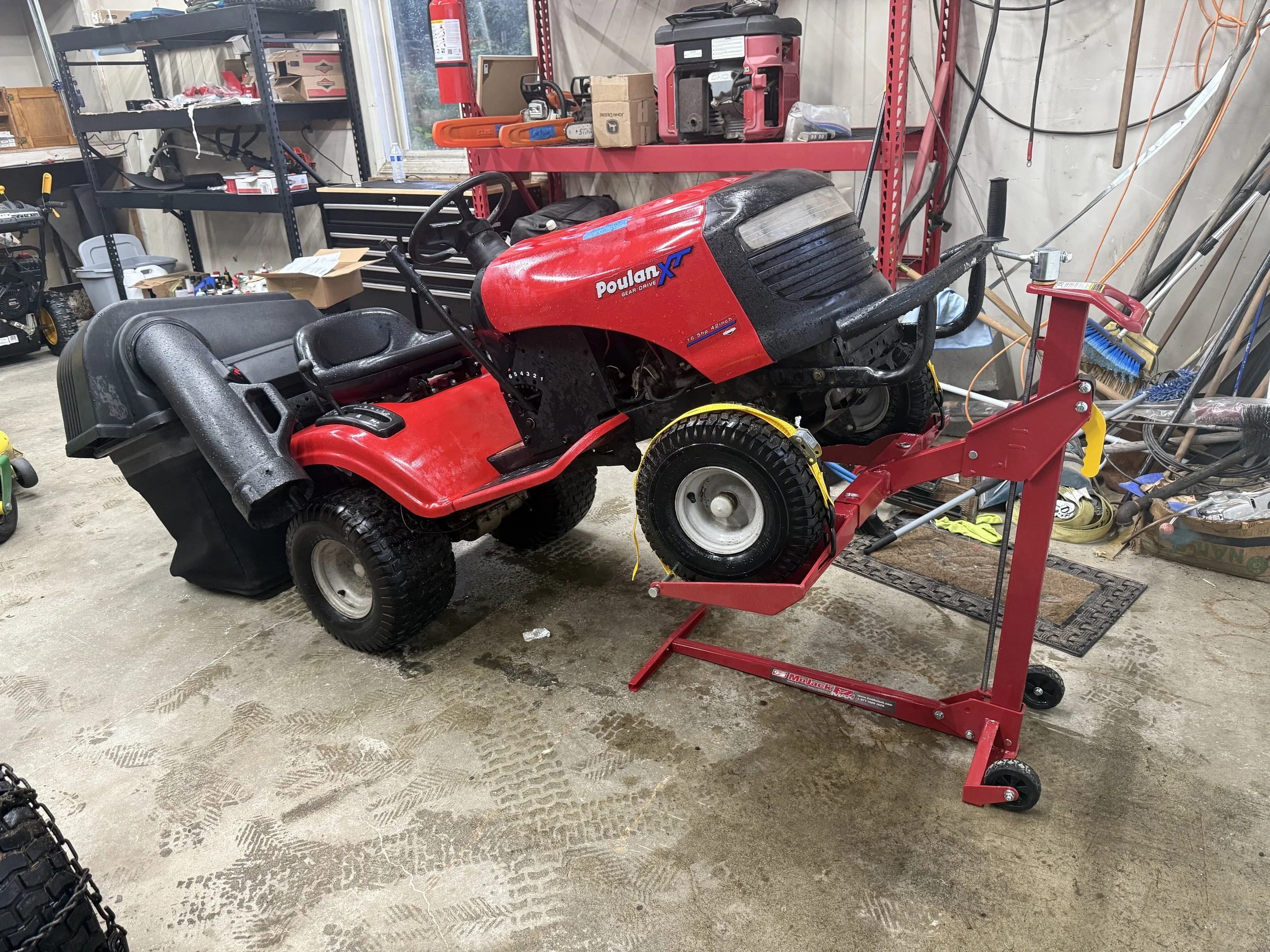 A red Poulan Pro riding lawn mower on a red stand with a front wheel removed, in a workshop with shelves, tools, and equipment around.