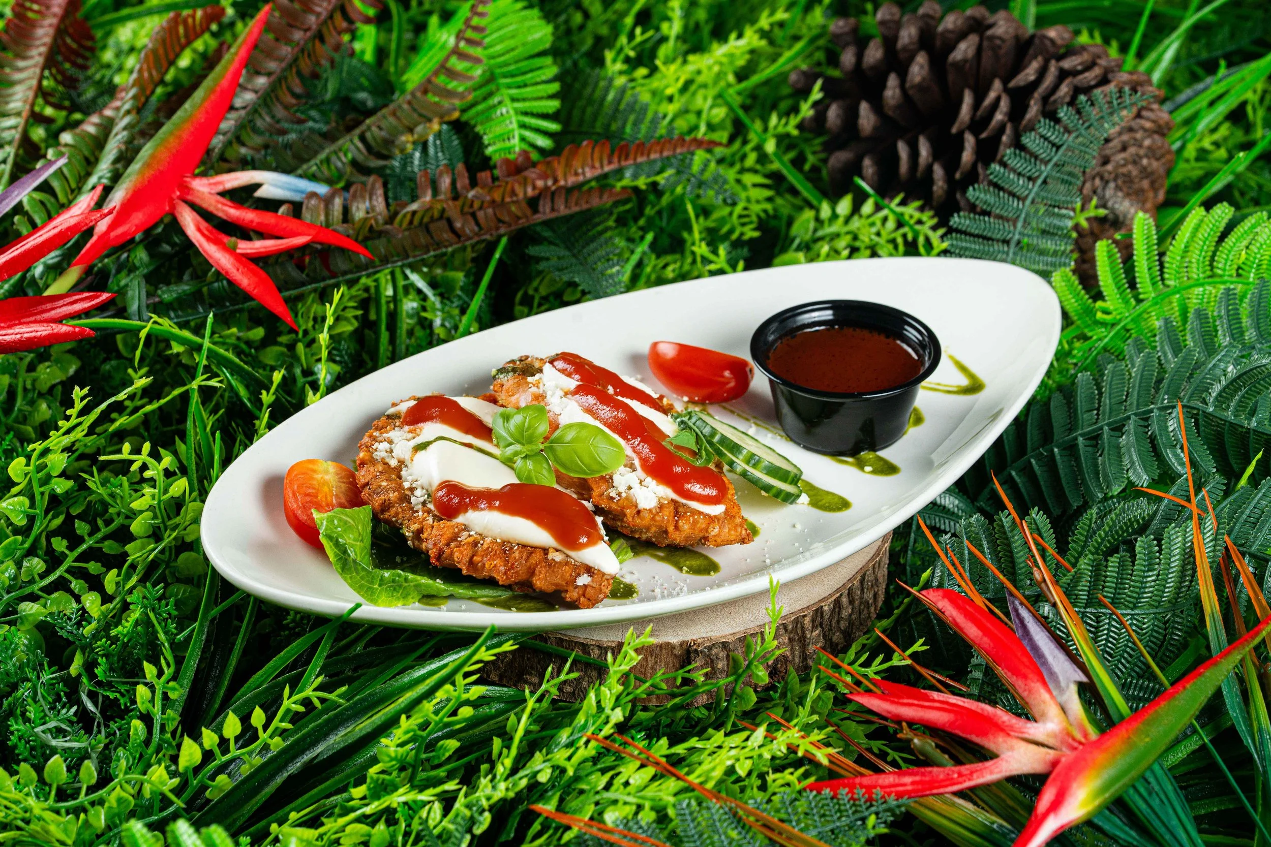 Fried chicken topped with mayonnaise, ketchup, and basil leaves, served on a white plate with cherry tomatoes, cucumber slices, and a side of barbecue sauce, placed on a round wooden slice among green tropical plants.