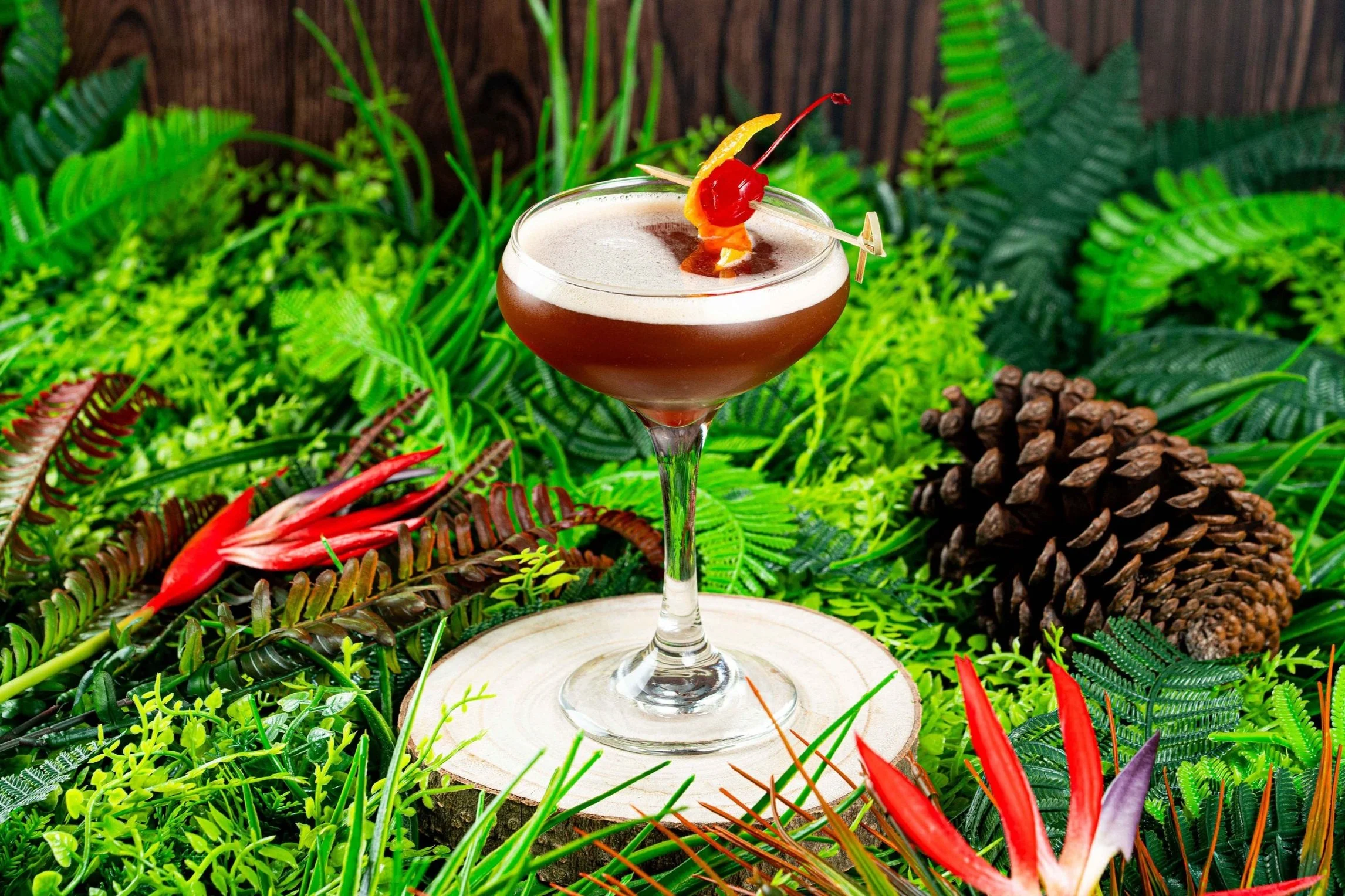 A cocktail glass with a chocolate-colored drink, garnished with a maraschino cherry and an orange twist, placed on a tree stump surrounded by green ferns, red flowers, and a pine cone.
