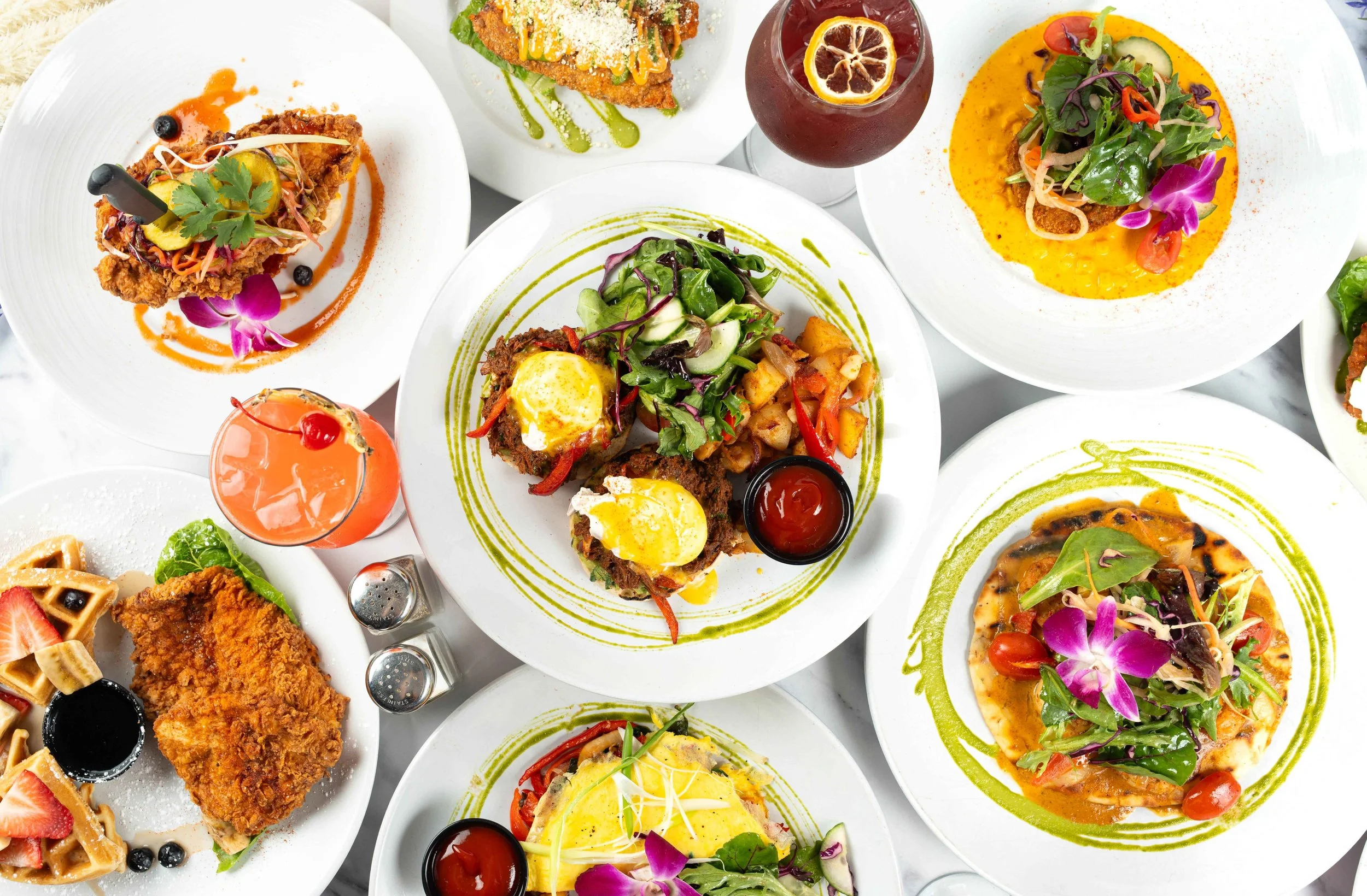 A variety of colorful dishes on white plates, including salads, pasta, fried chicken, and drinks, all garnished with flowers and vibrant sauces.