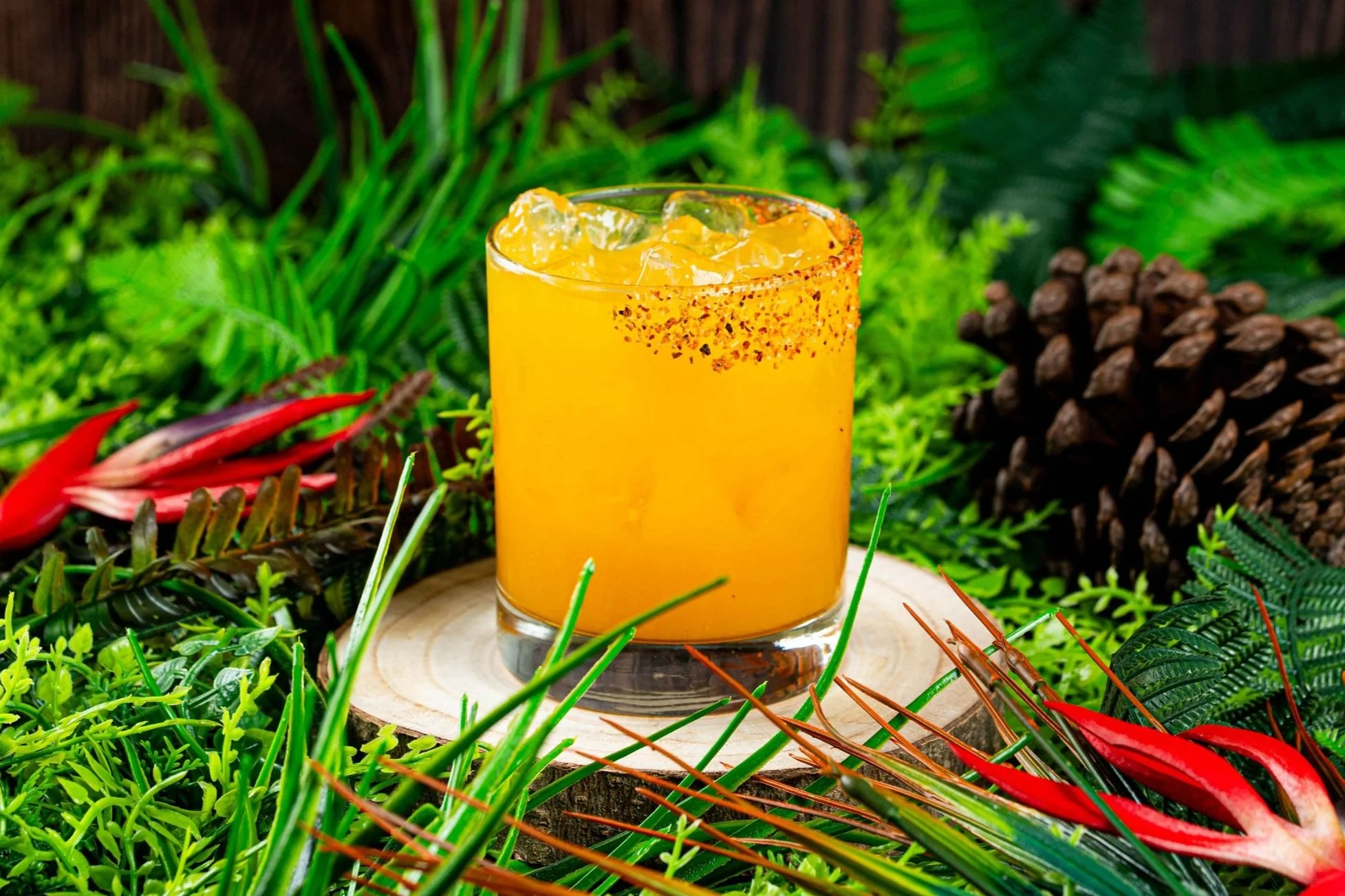 A glass of orange cocktail with ice and a rim of chili flakes, placed on a wooden slice surrounded by tropical plants and red flowers.