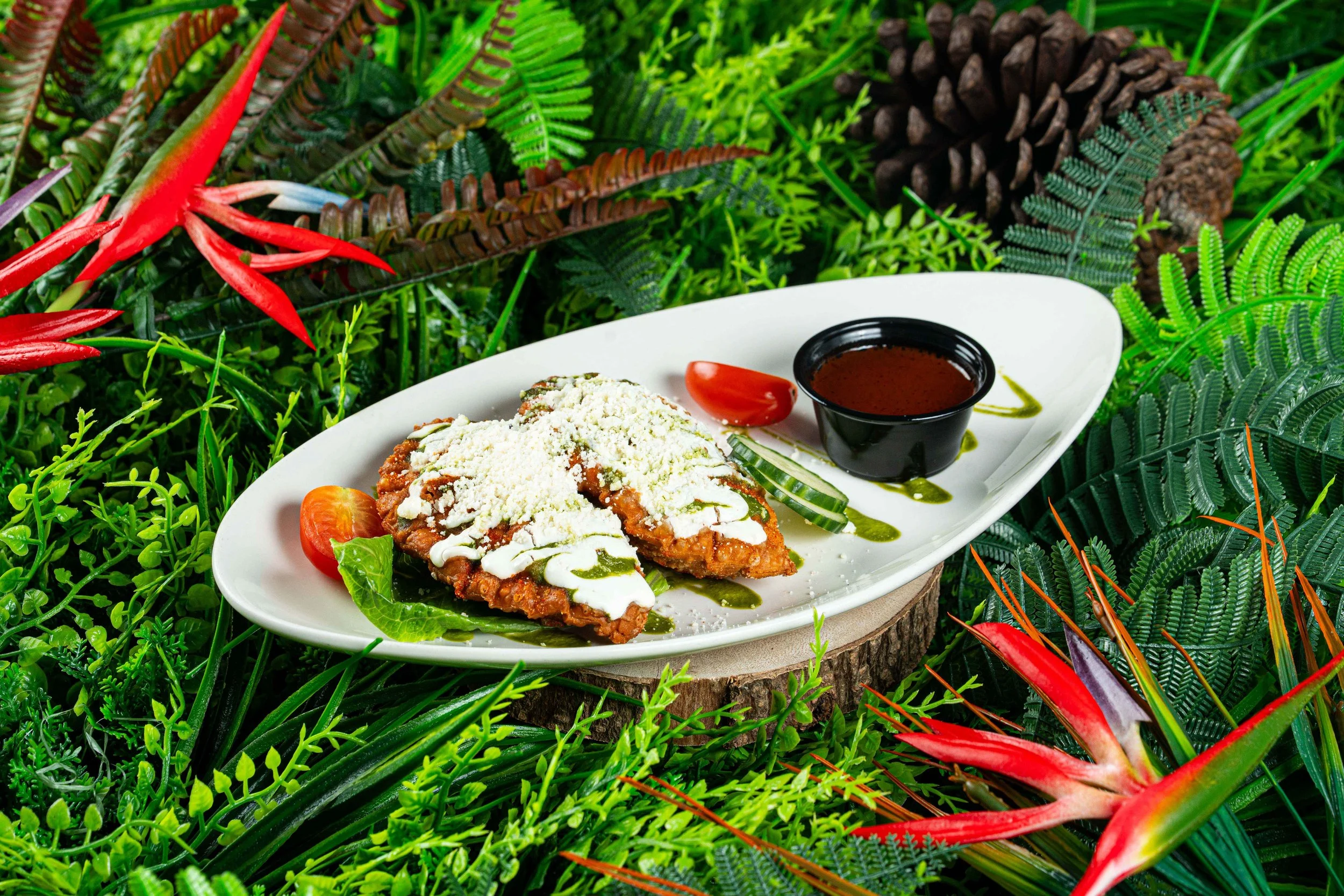 Plate of Mexican food with fried chicken topped with cheese, sauce, sliced cucumbers, cherry tomatoes, in a lush green jungle setting.