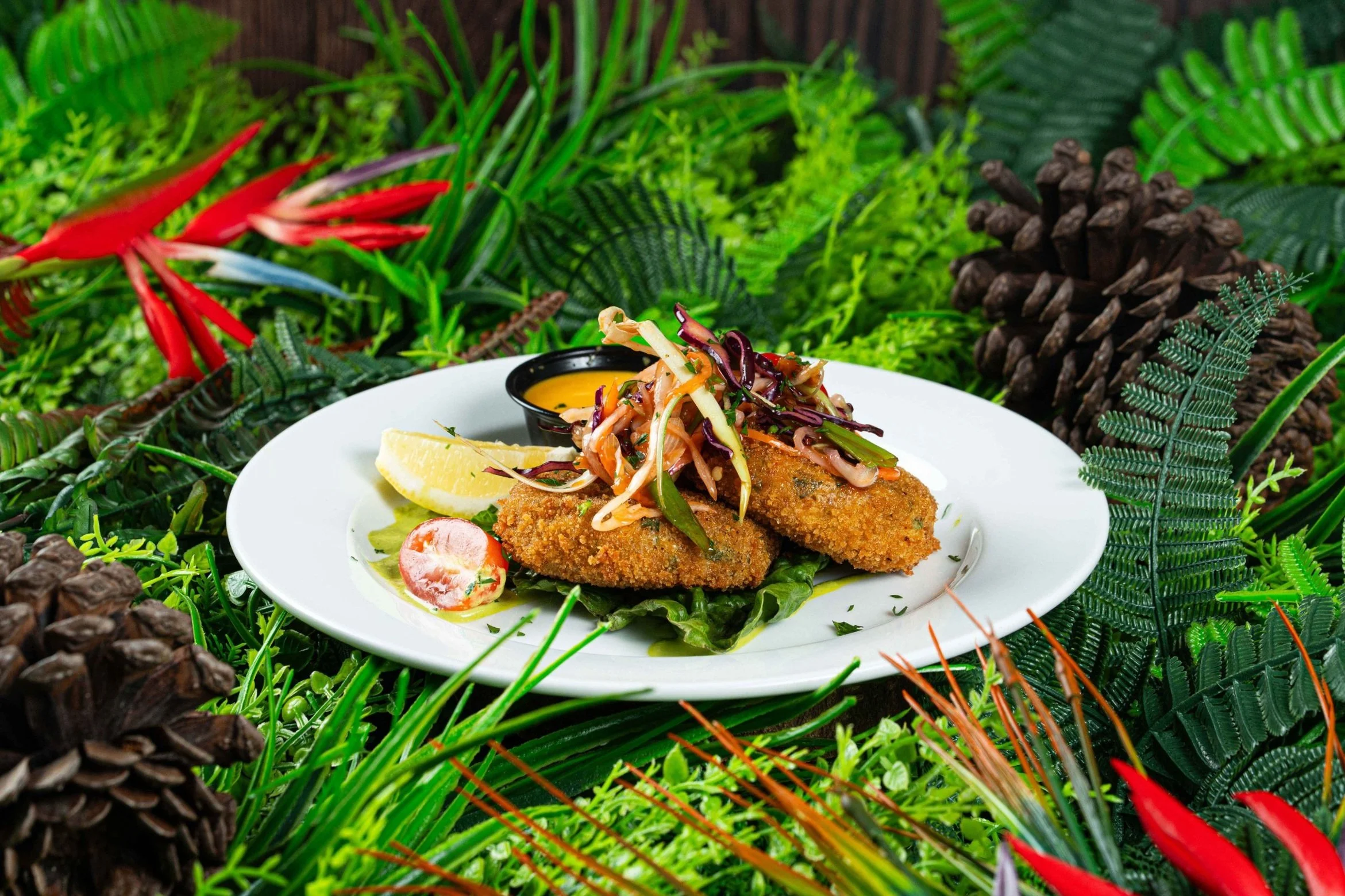 Fried fish fillets with coleslaw, lemon wedge, cherry tomato, and dipping sauce on a white plate, surrounded by a lush green tropical foliage background.