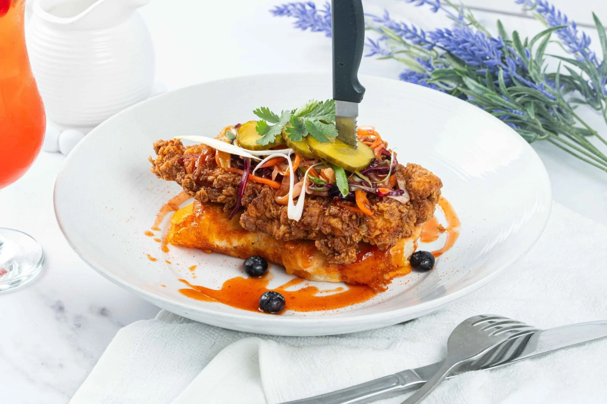 Fried chicken served on a bed of pasta with sauce, topped with pickles, herbs, and vegetables, garnished with black berries.