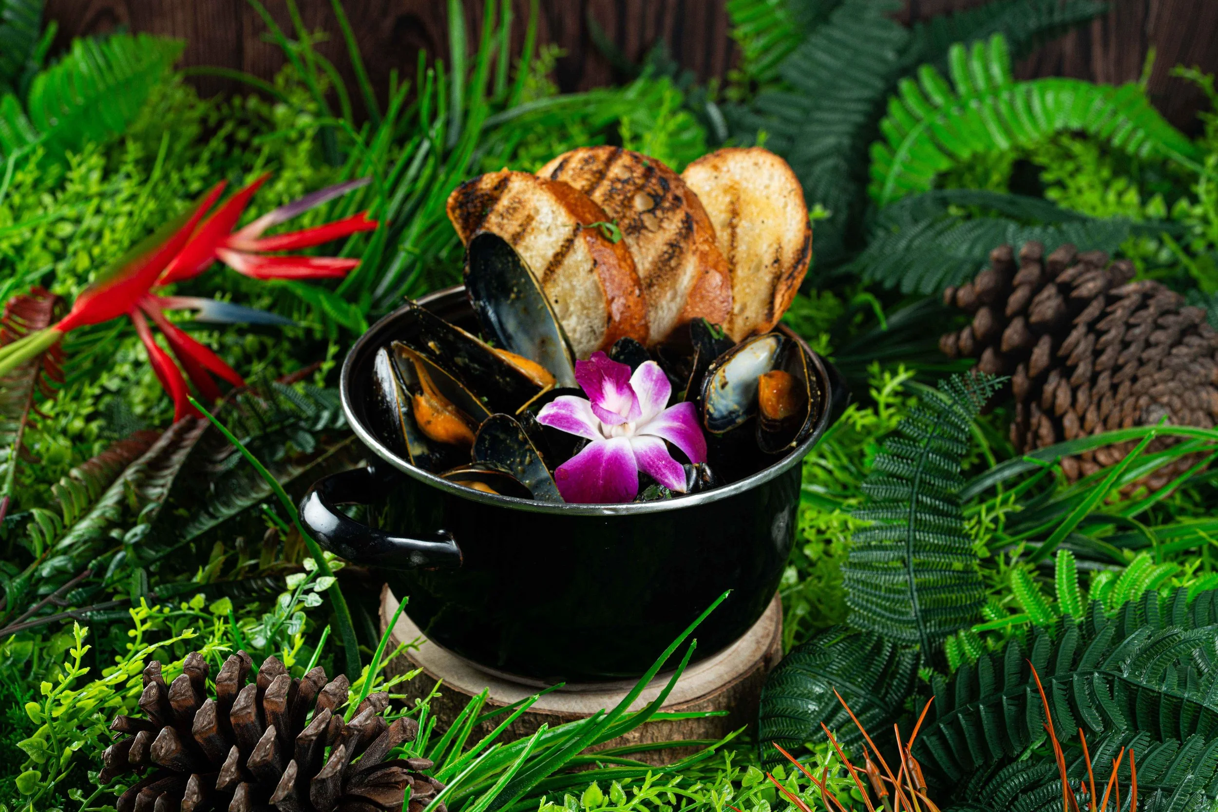 A black cup of steamed mussels garnished with a purple flower, served with toasted bread slices, surrounded by lush green tropical plants and pinecones.
