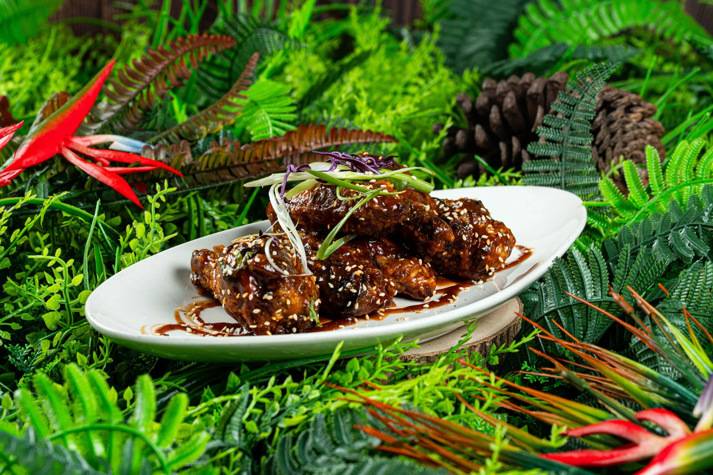 Plate of glazed chicken wings garnished with sliced green onions on a white dish, surrounded by tropical leaves and flowers.