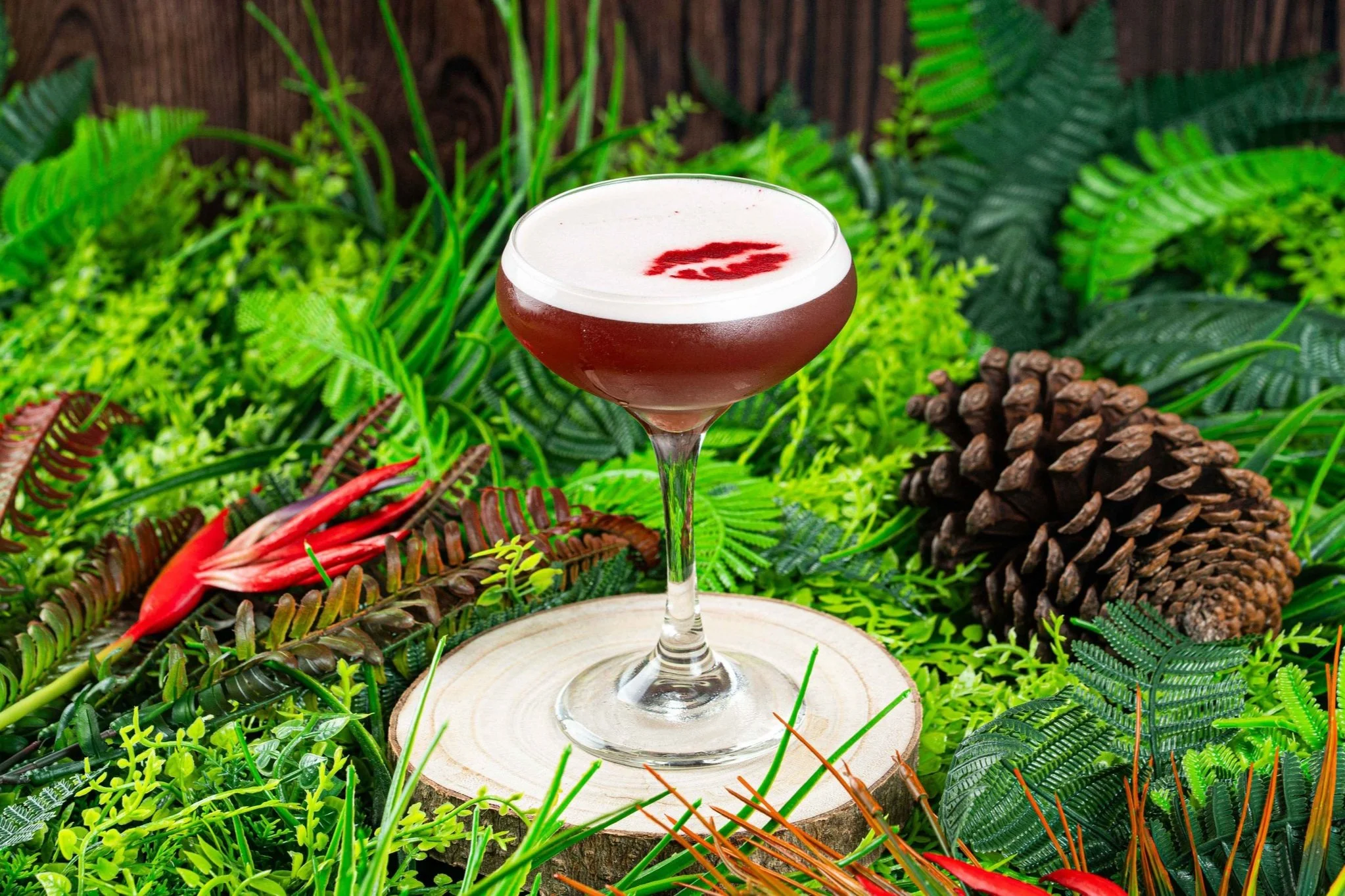 A cocktail in a stemmed glass with a frothy top and red garnish, placed on a tree slice, surrounded by green greenery and pinecones.