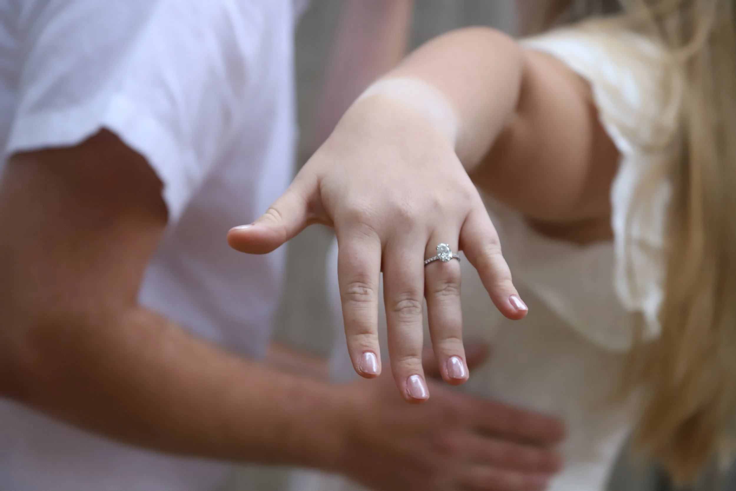 Close-up of a woman's hand showing an engagement ring, with a blurred background of a man and woman.