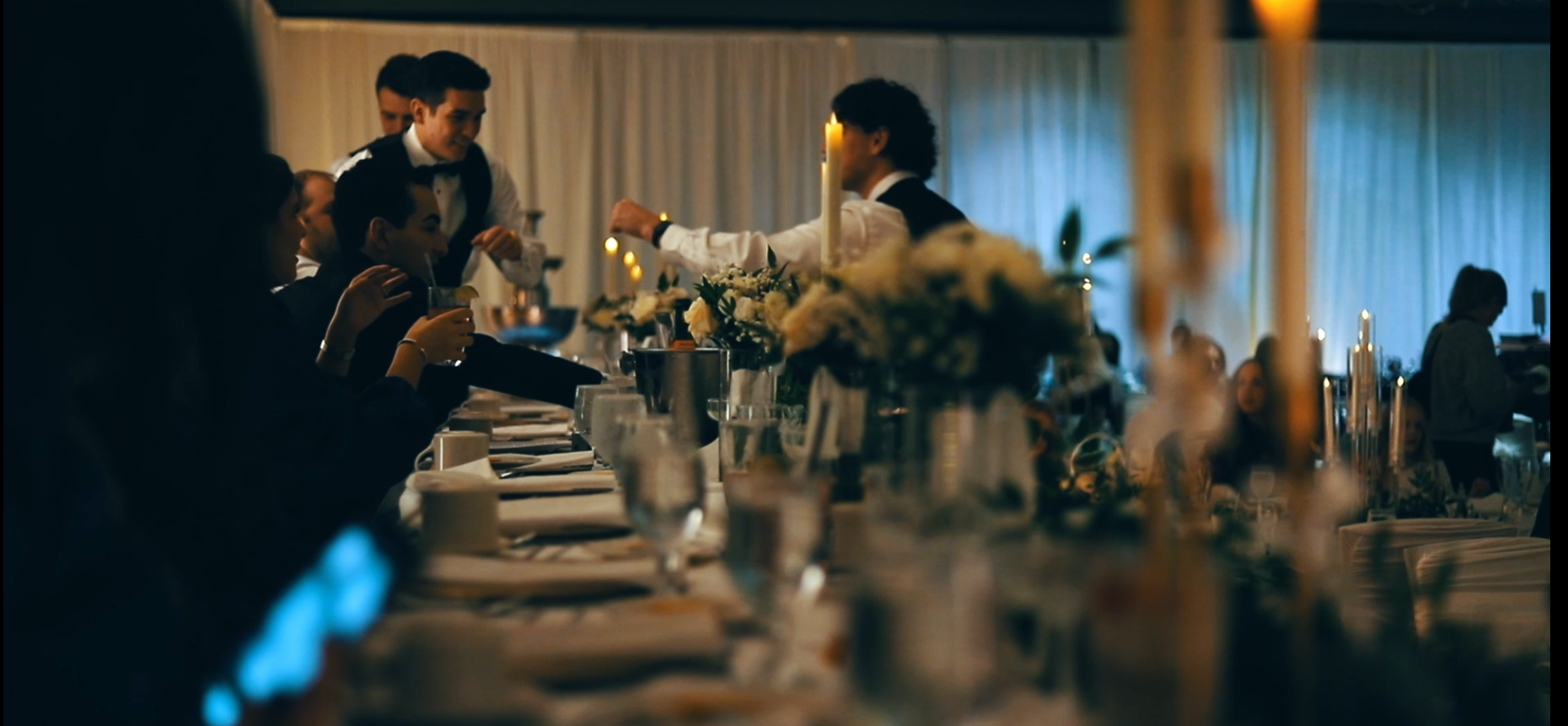 People sitting and standing around a decorated dining table with candles and flowers at an elegant event or celebration.