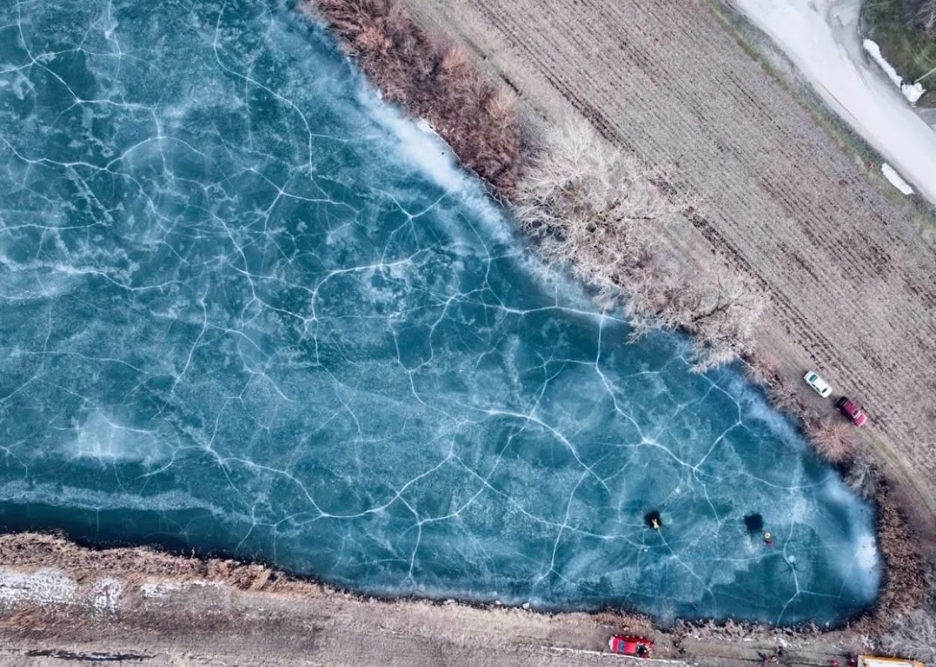 An aerial view of a frozen pond with visible cracks, surrounded by trees, a dirt field, and parked cars.