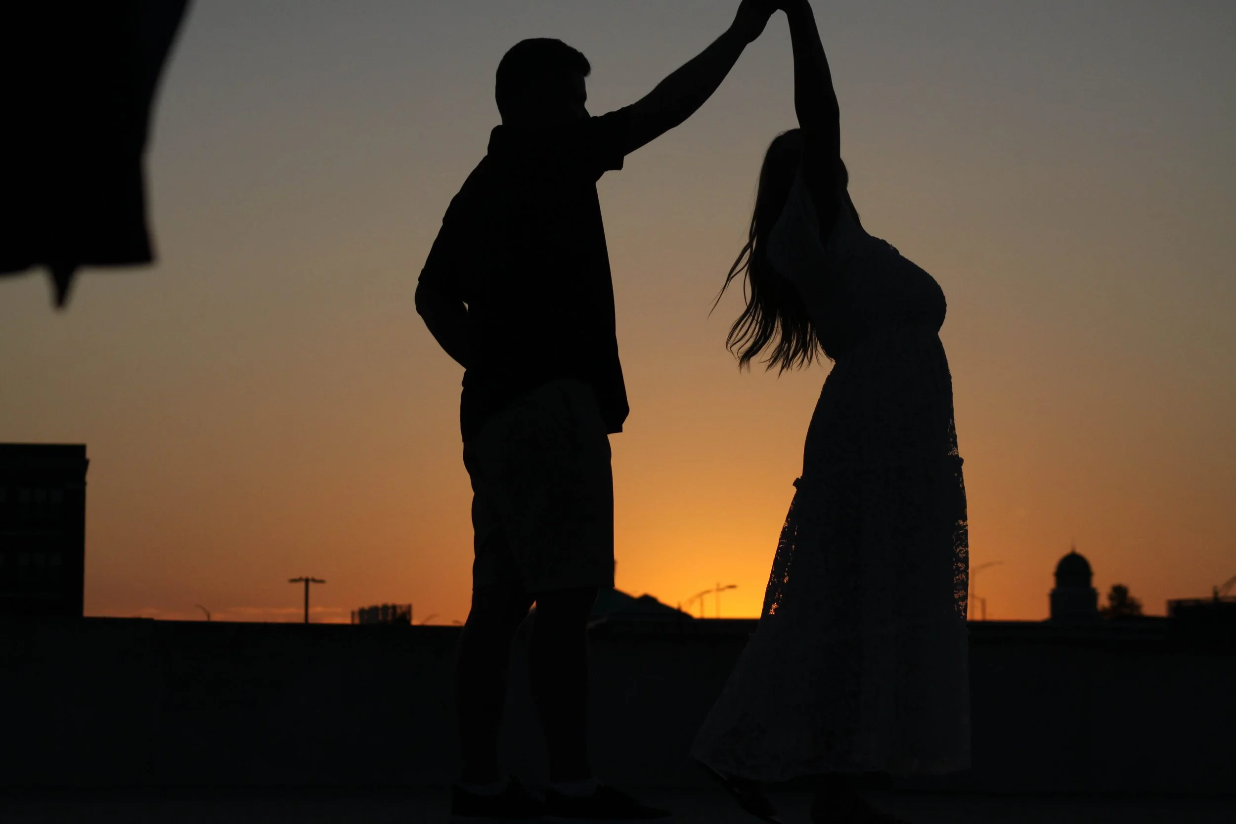 Silhouette of a man and woman dancing at sunset on a rooftop, city skyline in the background.