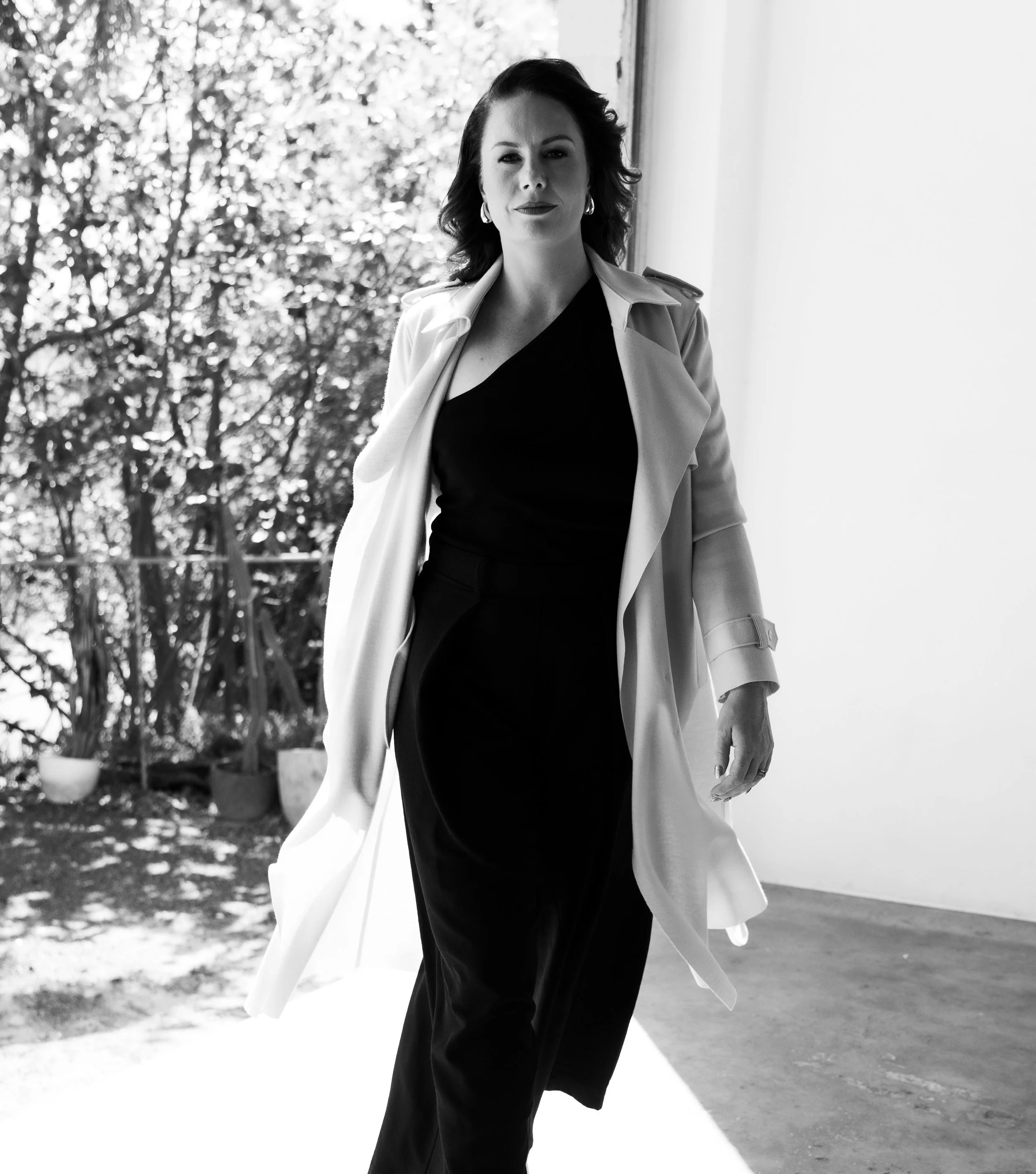 A woman with dark shoulder-length hair wearing a light-colored trench coat, black top, and black pants, walking indoors with natural light coming from the right, and a background of greenery visible through a large window.
