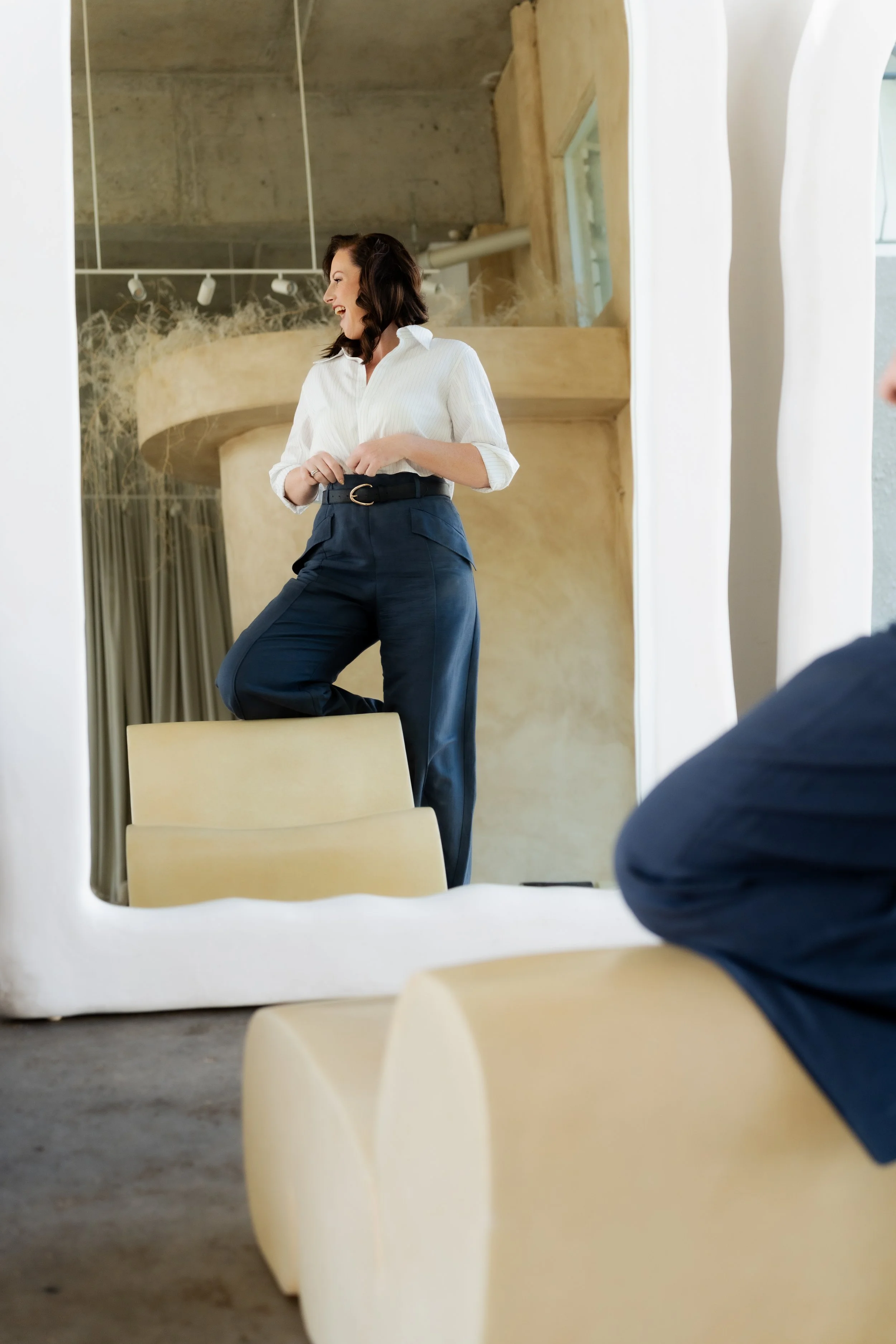 A woman with dark hair, wearing a white shirt and navy trousers, is standing on a chair in front of a large mirror. She is smiling and looking to her left, with her hands clasped in front of her. This appears to be a modern, stylish interior space with a concrete ceiling and neutral-colored decor.