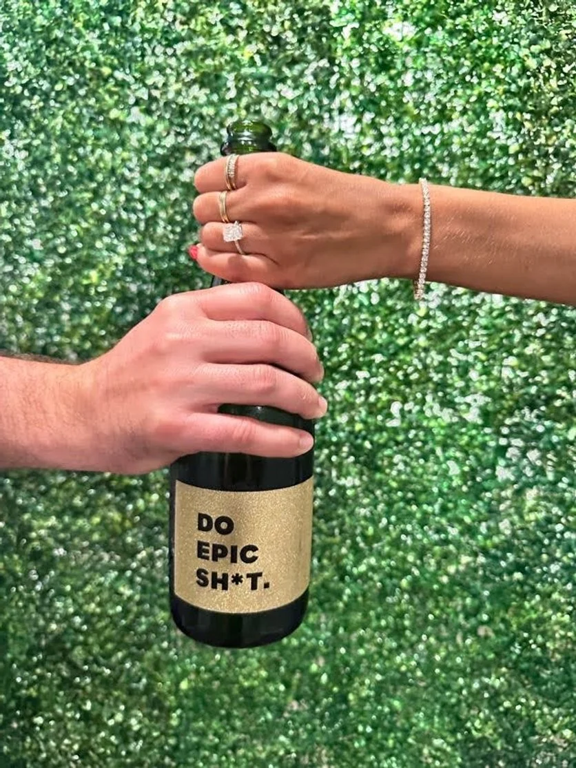 Two hands, one with multiple rings and a bracelet, holding a bottle of champagne or sparkling wine. The bottle has a beige label with bold black text that reads 'DO EPIC SH*T.' The background is a green, glittery, textured surface.