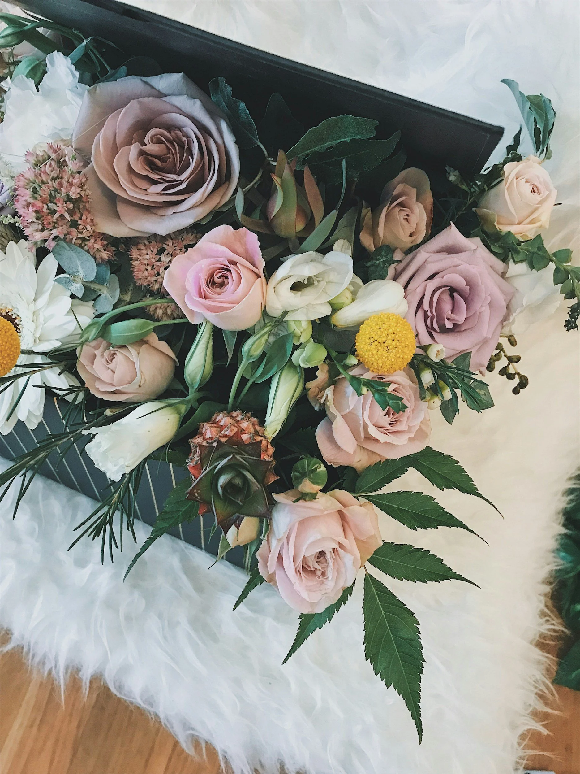 Arrangement of pink, white, and lavender roses with greenery in a black box, placed on a white furry surface.