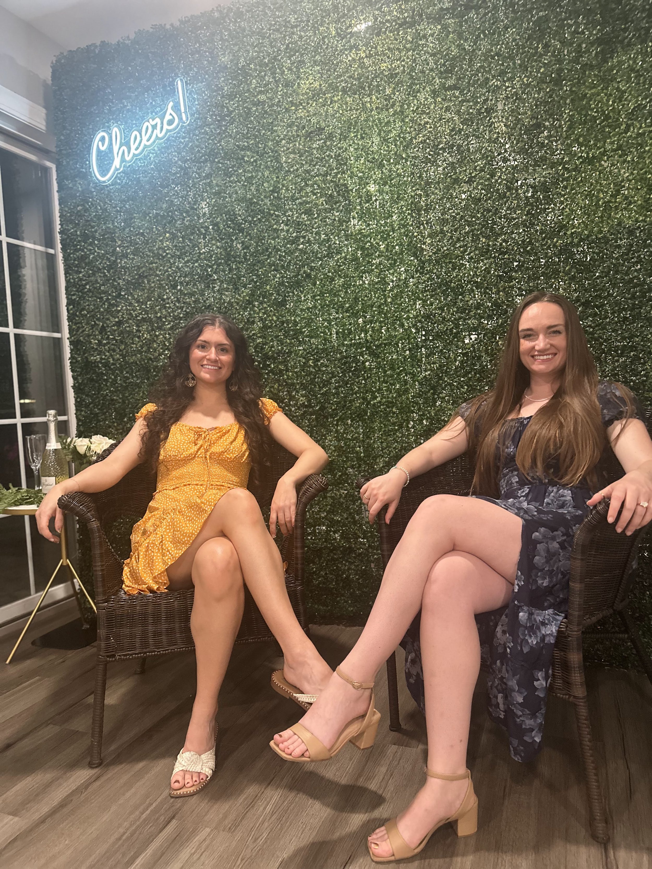Two young women sitting in wicker chairs in front of a green hedge wall with a neon sign that says 'Cheers!'. One woman is wearing a yellow dress with white sandals, and the other is wearing a dark floral dress with beige heels. They are smiling and relaxed.