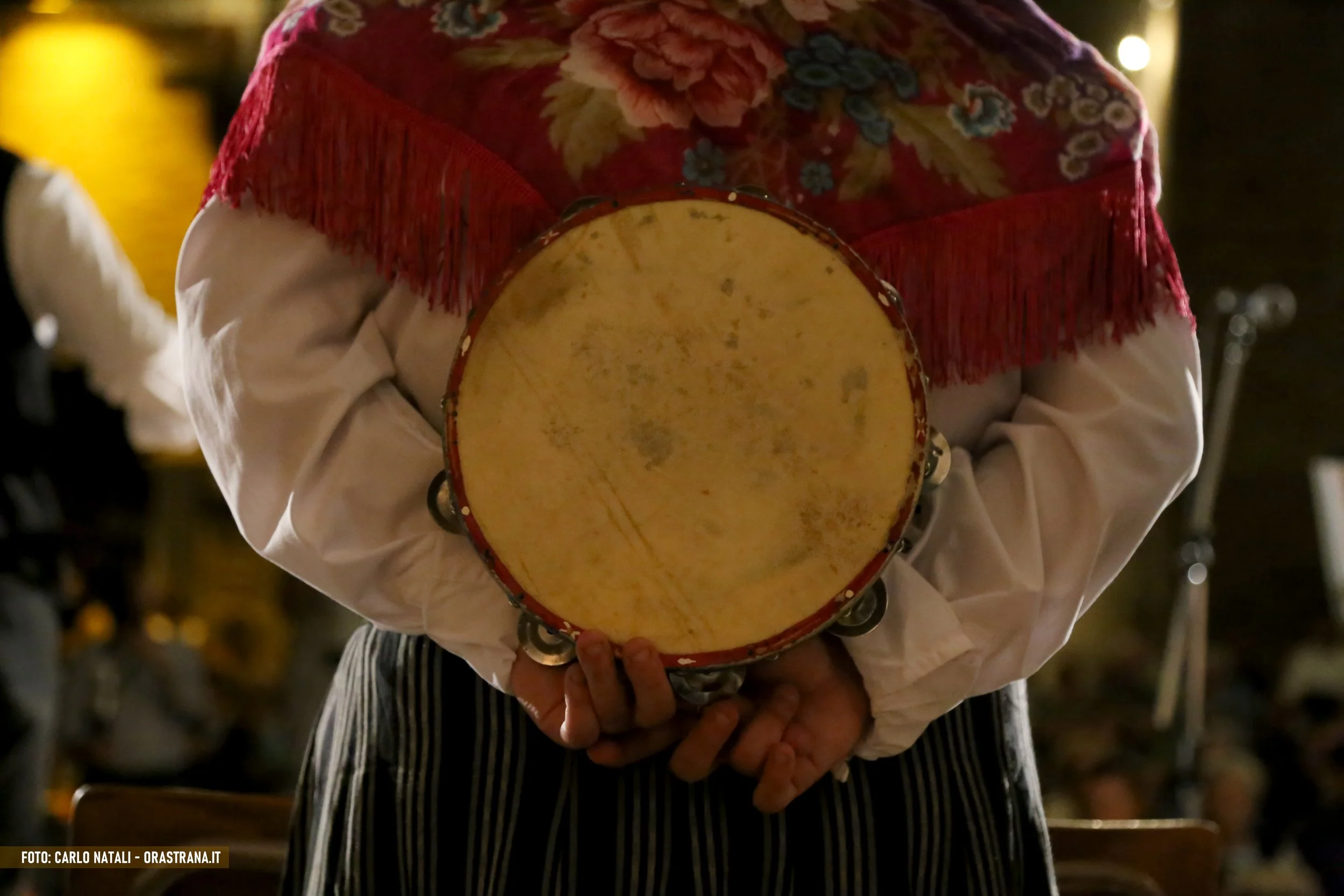 Person holding a tambourine, dressed in traditional clothing with a floral shawl.