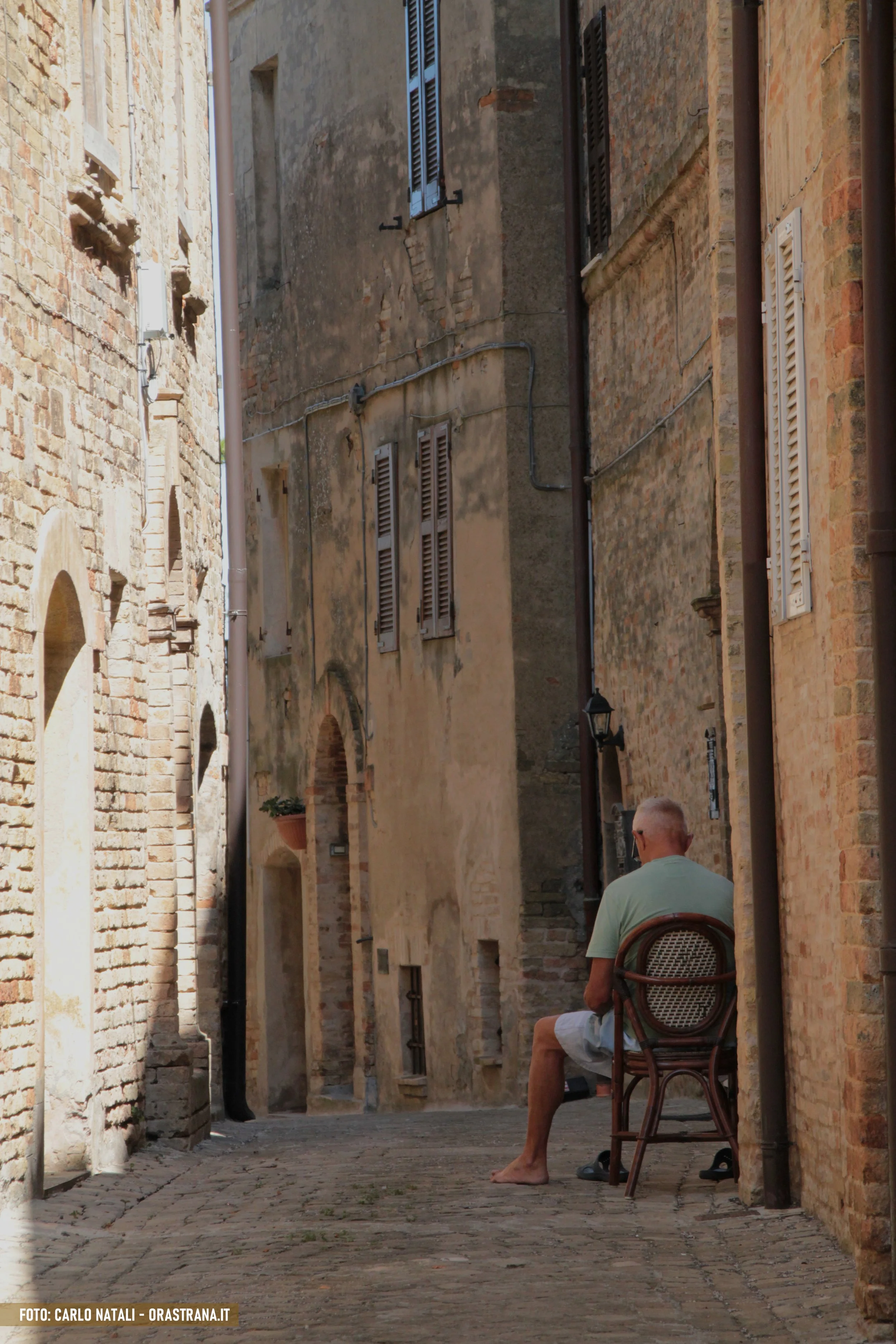 An elderly man with gray hair and sunglasses sitting on a wooden chair in a narrow European alleyway, surrounded by old brick and stucco buildings with shutters and small windows.