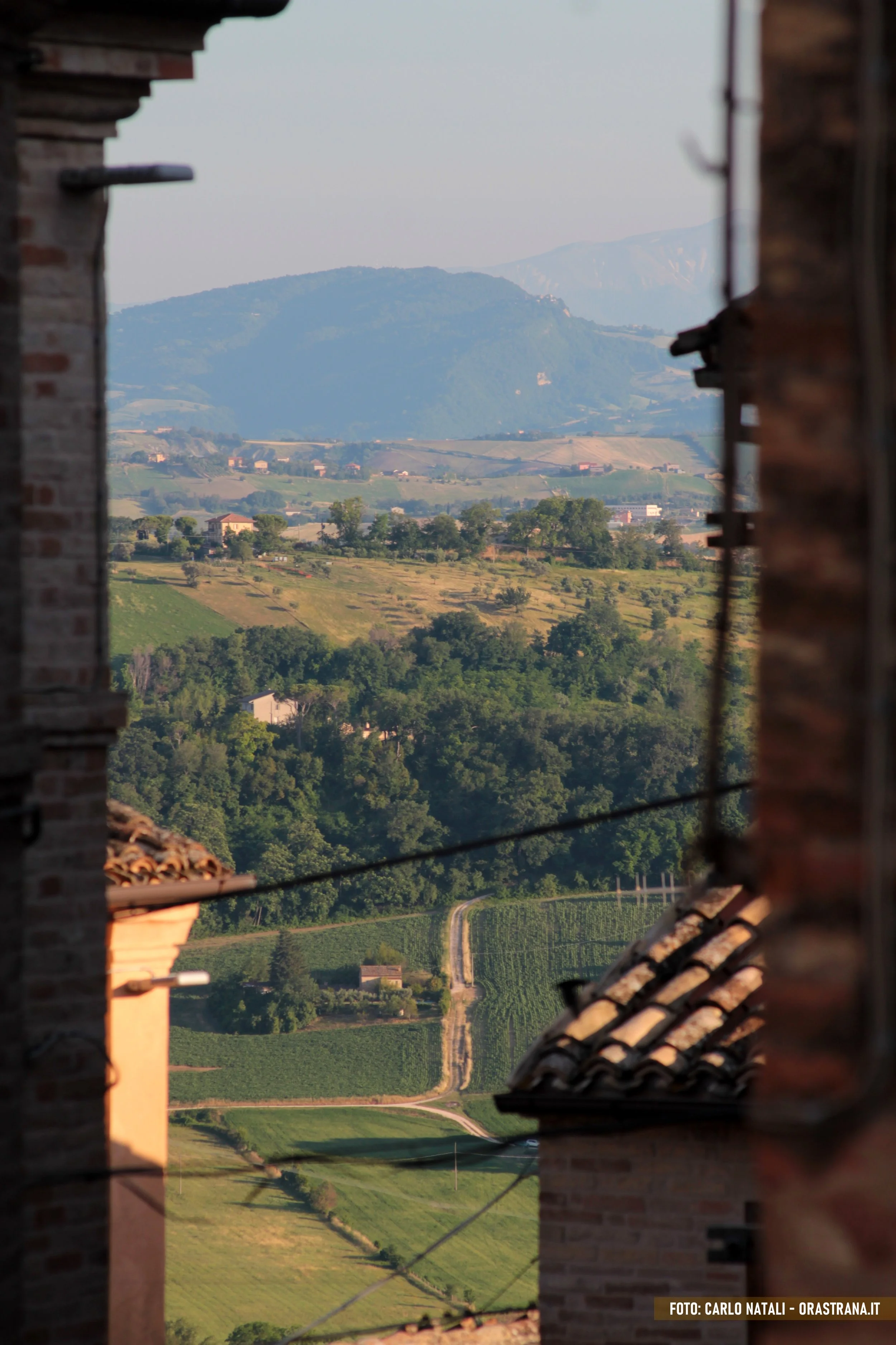 A scenic view of lush green hills, vineyards, and distant mountains seen through a framed opening between brick buildings.