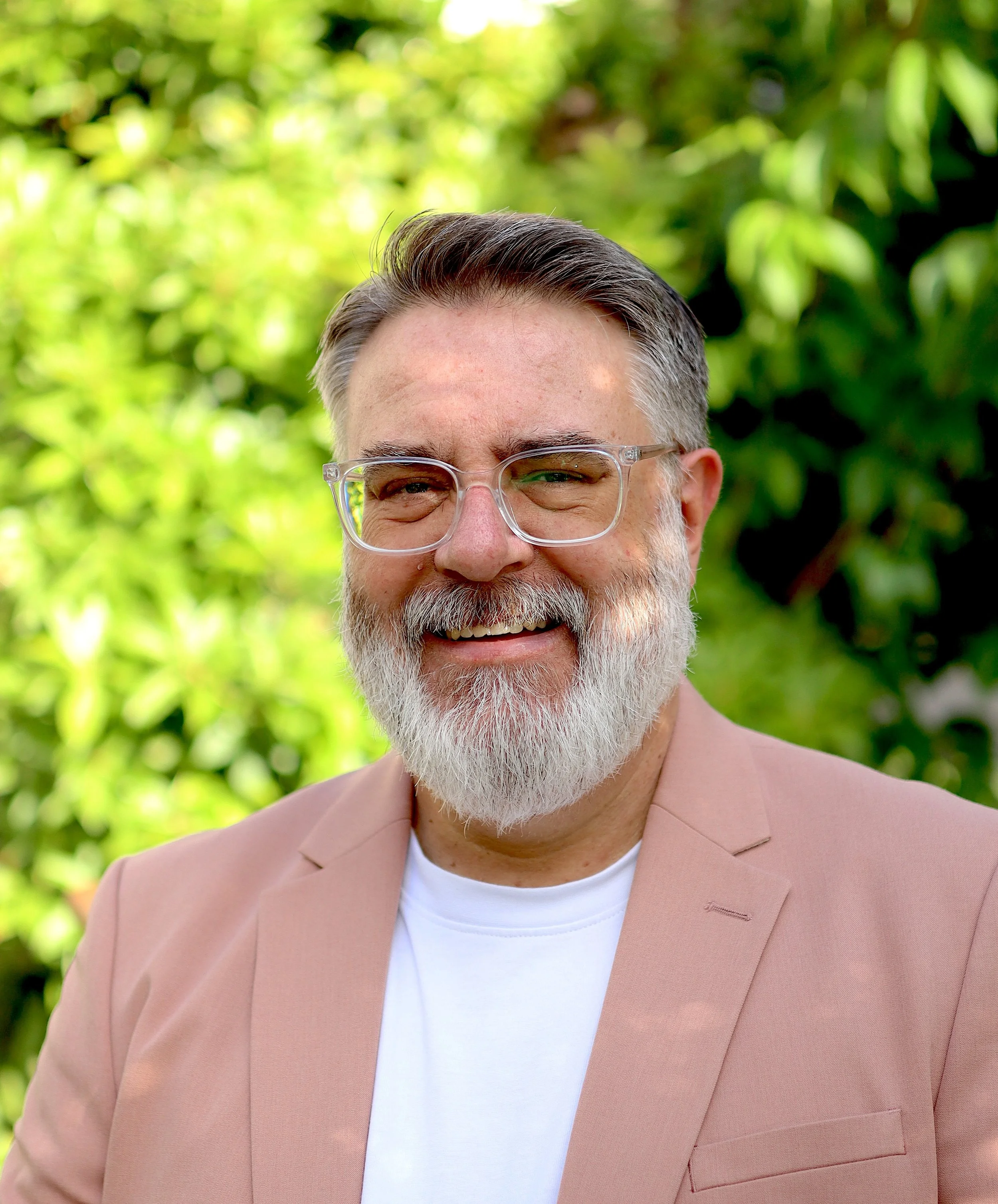 A smiling white man with a grey beard wearing a white T-shirt and salmon blazer