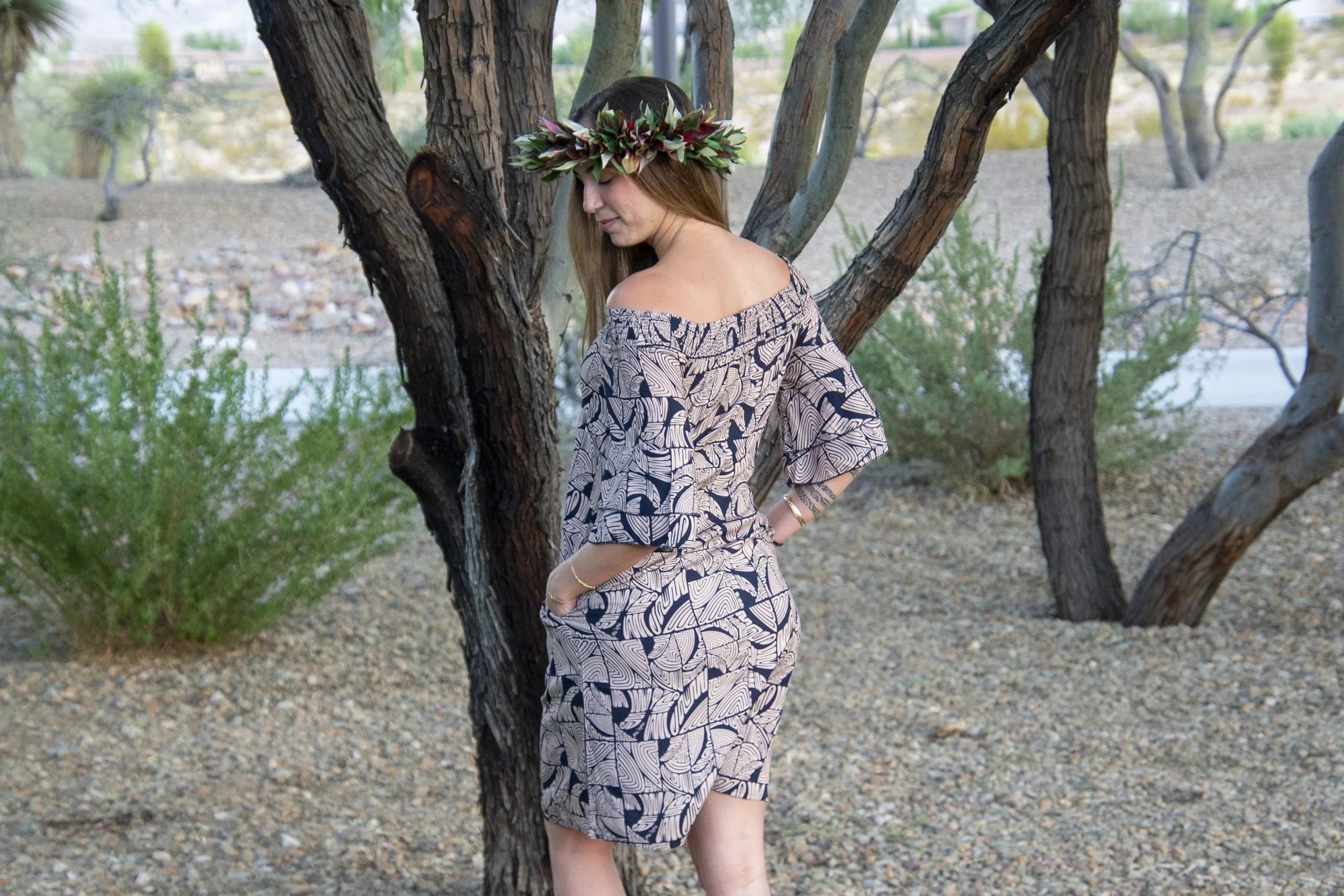 A woman wearing a patterned off-the-shoulder dress and a floral crown standing outdoors near a tree in a desert-like setting.