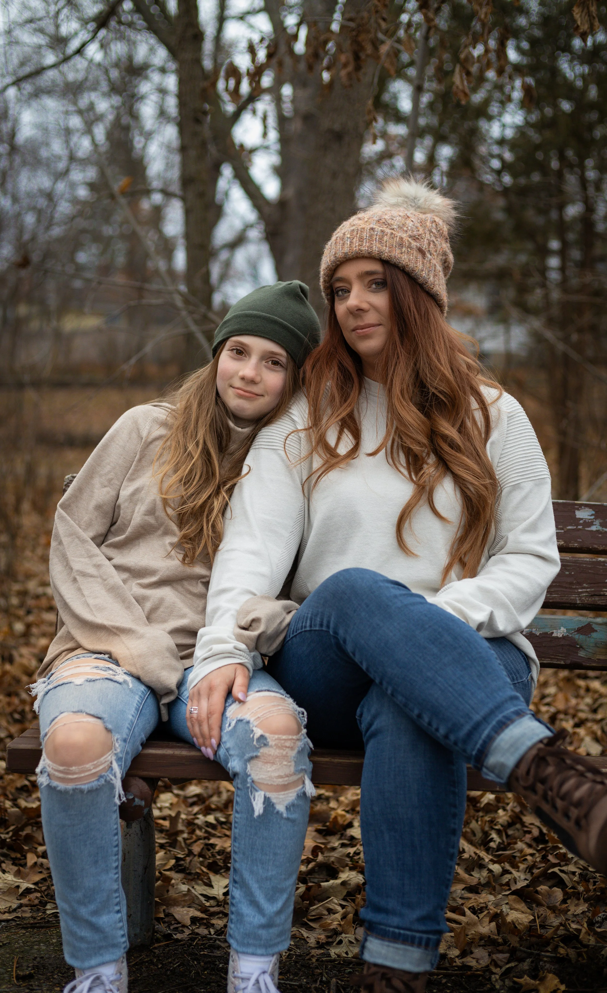 A woman and a girl sitting close together on a wooden park bench surrounded by fallen autumn leaves, wearing casual clothes and beanies, with a blurred background of trees.