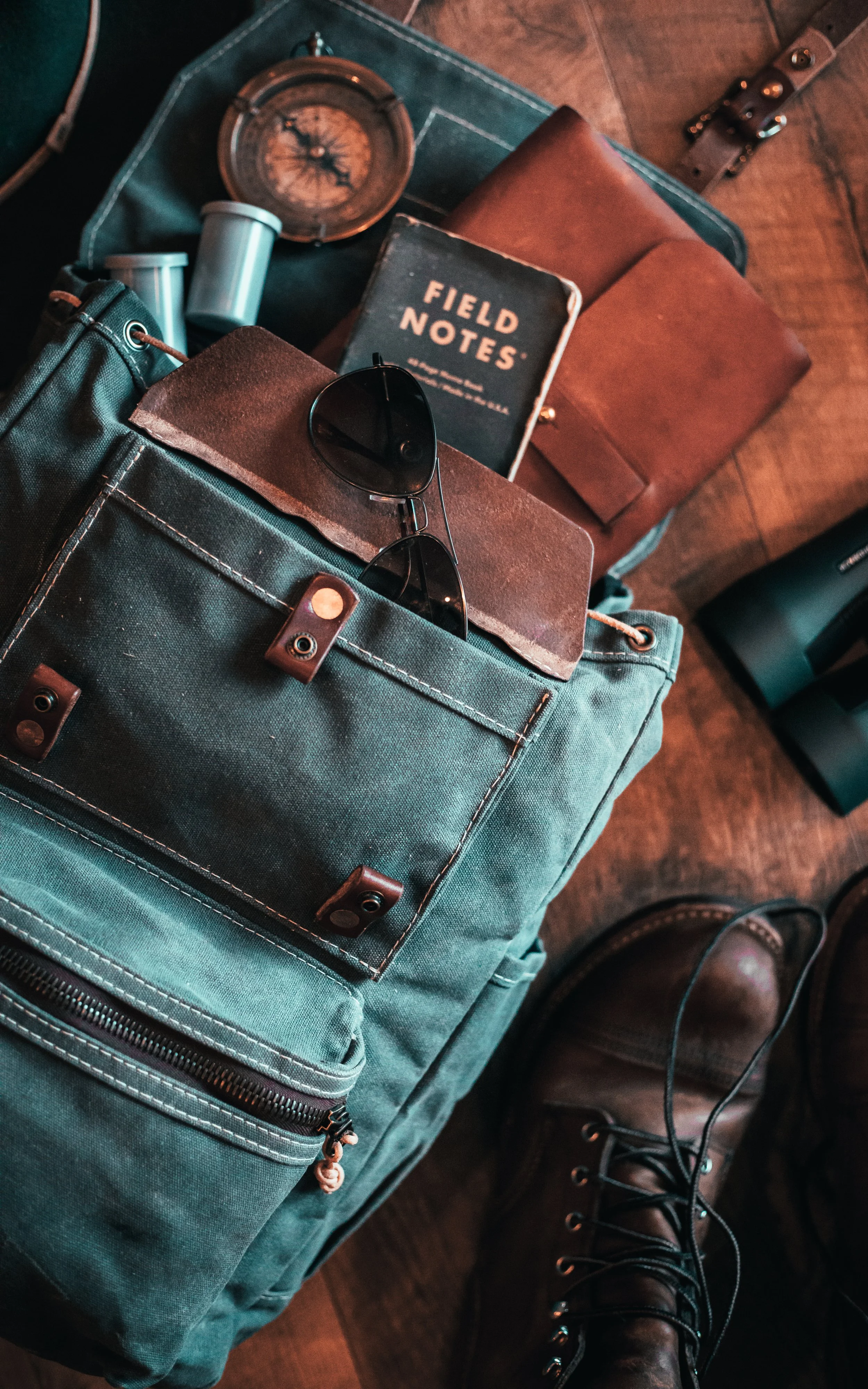Travel backpack with sunglasses, field notes journal, leather wallet, compass, and leather shoes on wooden floor.