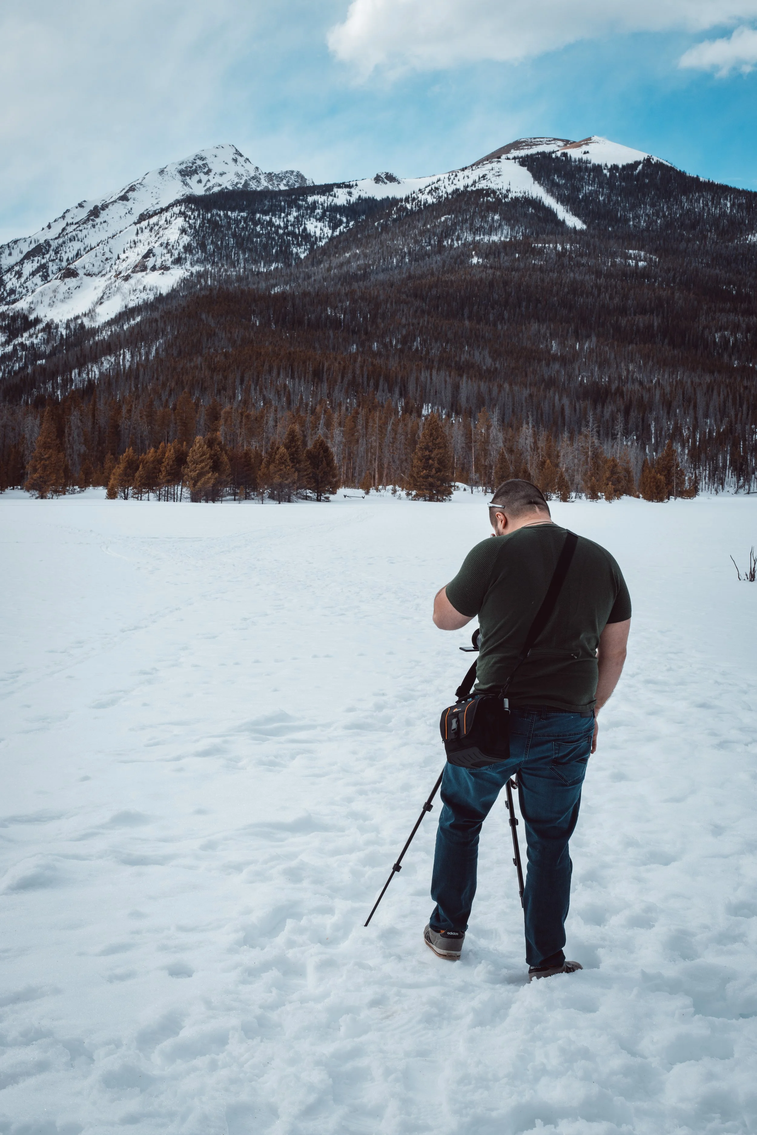 Man standing in snowy landscape with camera and tripod, mountains with snow and trees in the background