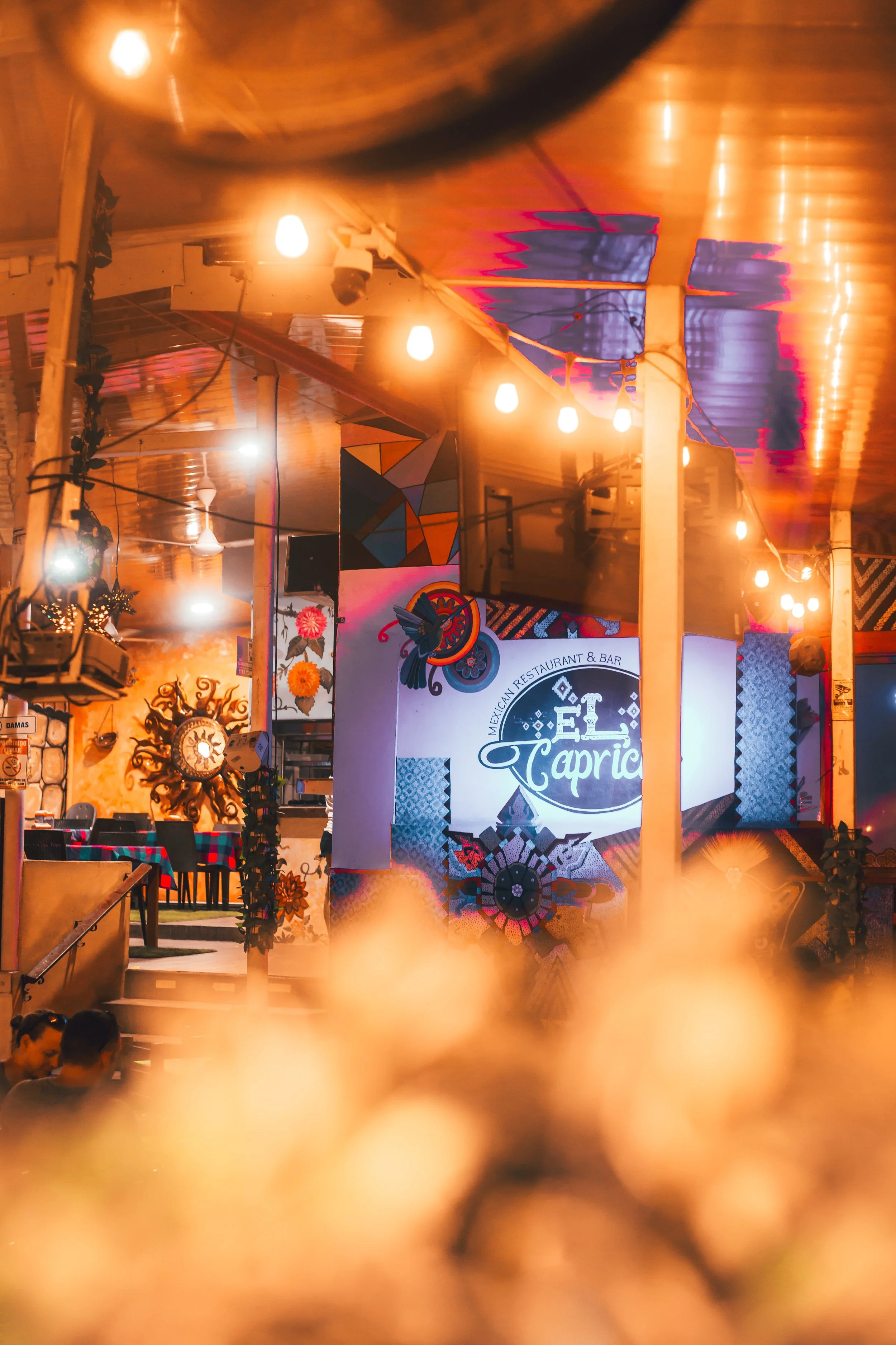 Interior of a Mexican restaurant and bar called El Caprice, decorated vibrant with wall art, hanging lights, and tables.