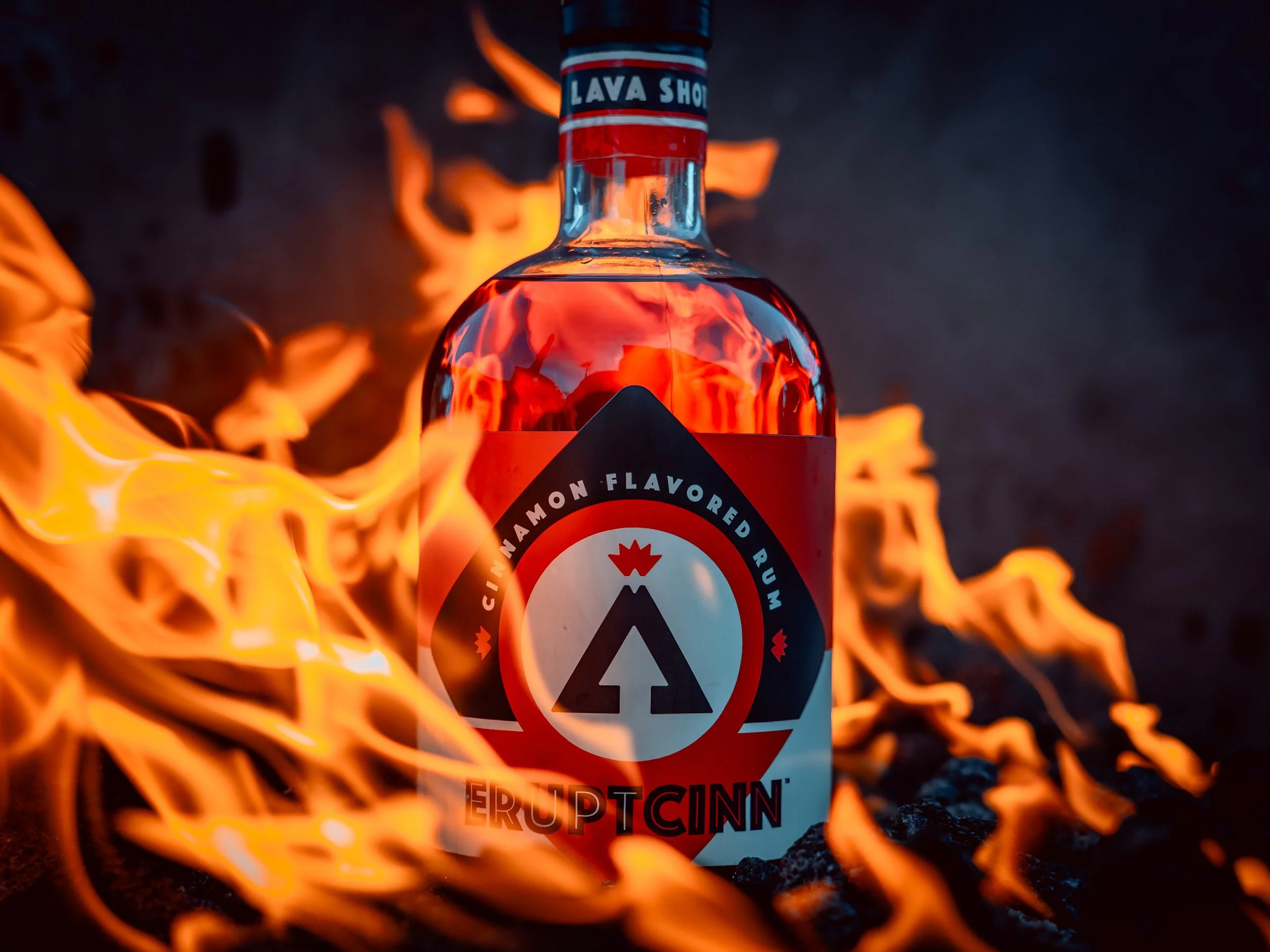 A bottle of EruptCinn cinnamon flavored rum surrounded by flames, with a black, red, white, and blue label featuring a volcano logo and the words "Cinnamon Flavored Rum" and "EruptCinn".