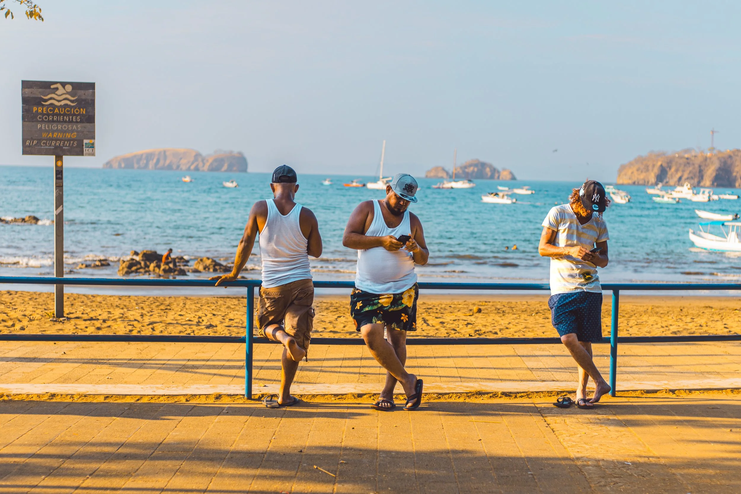 Three men standing by a blue railing on a sandy beach, looking at their phones, with boats in the water and islands in the background.