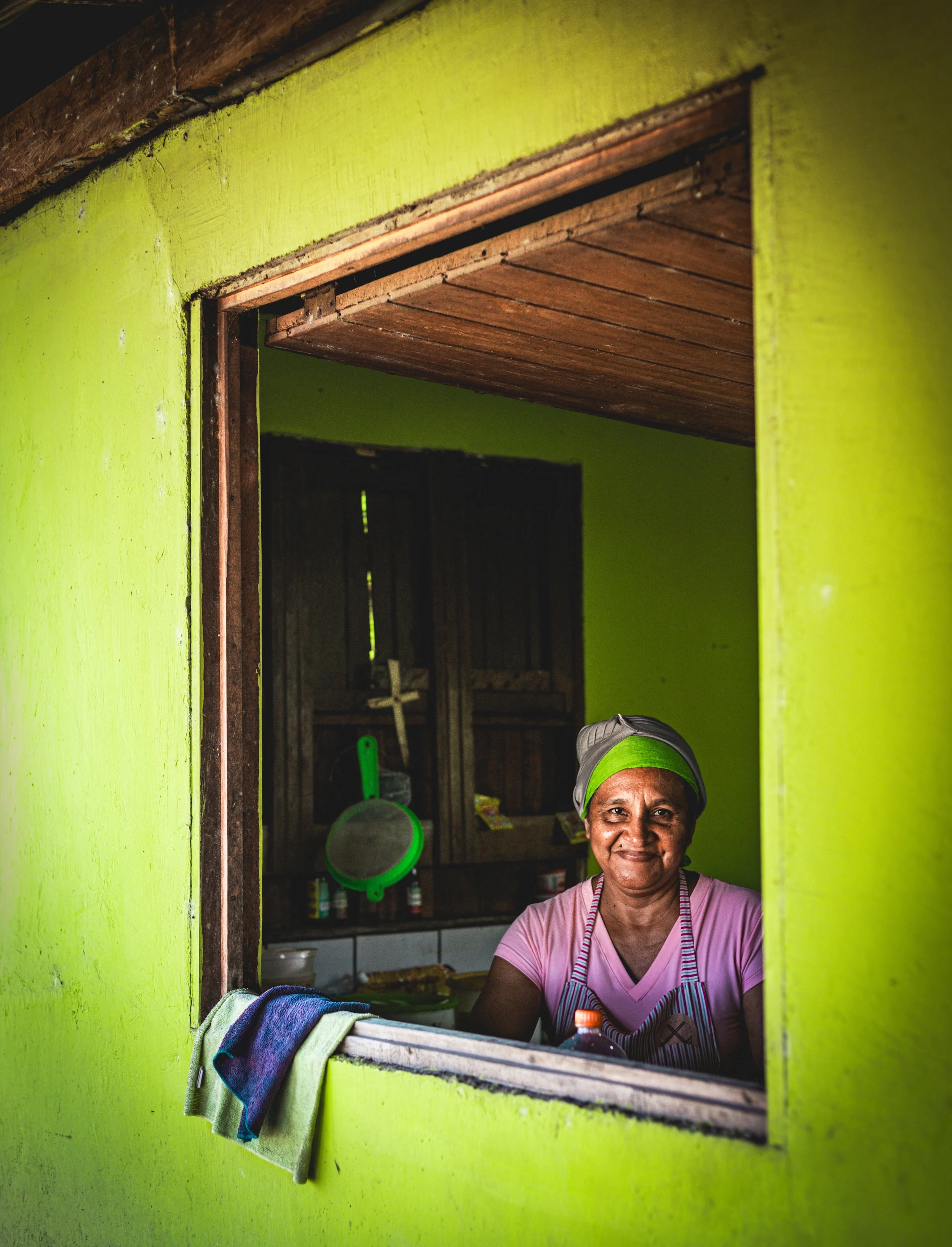 A woman in a pink shirt and green headscarf standing inside a kitchen, seen through a window with bright green walls, with a towel hanging on the window ledge.
