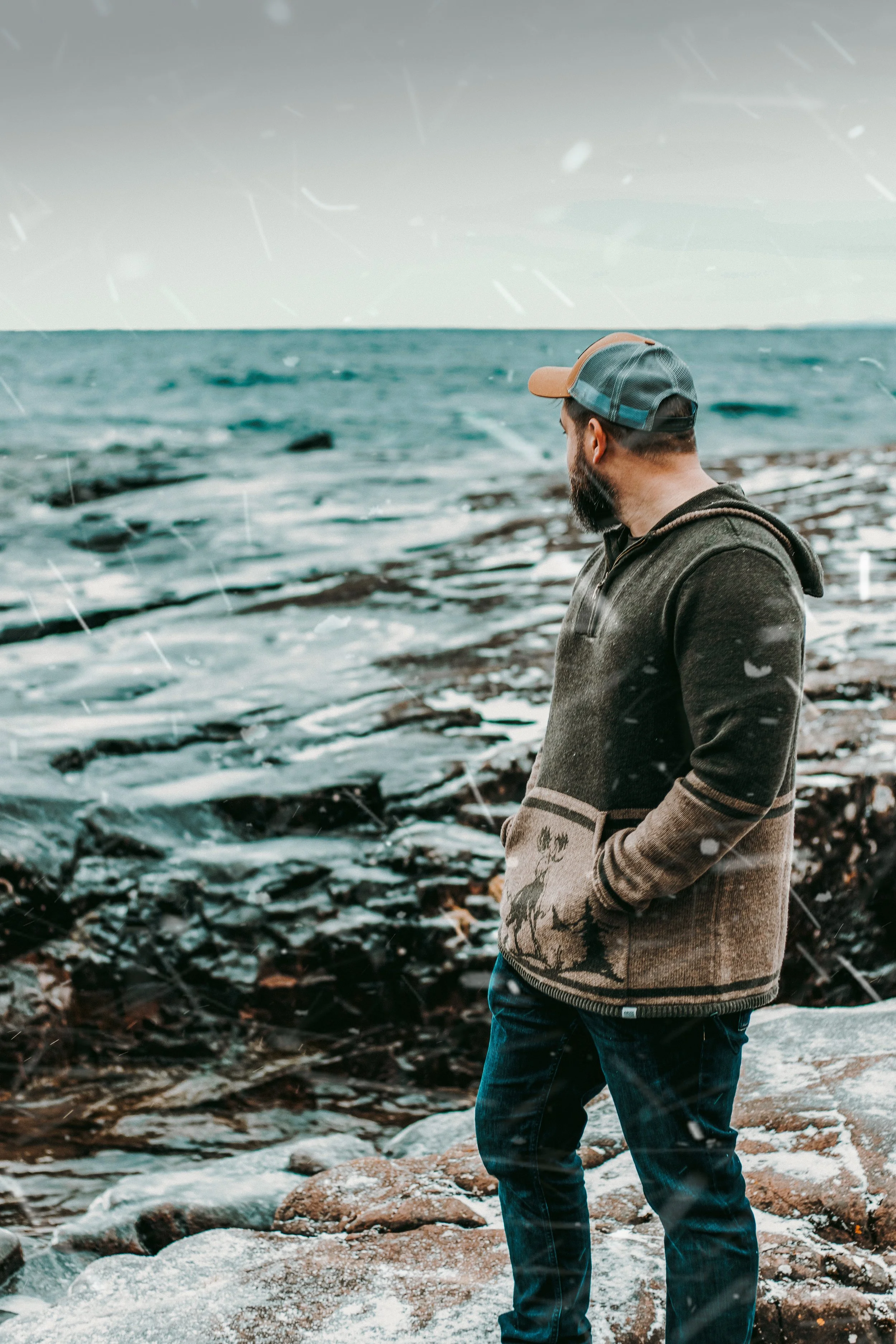 A man with a beard wearing a gray hoodie with reindeer pattern and a baseball cap, standing on rocky shoreline, looking at the ocean with waves during snowy weather.