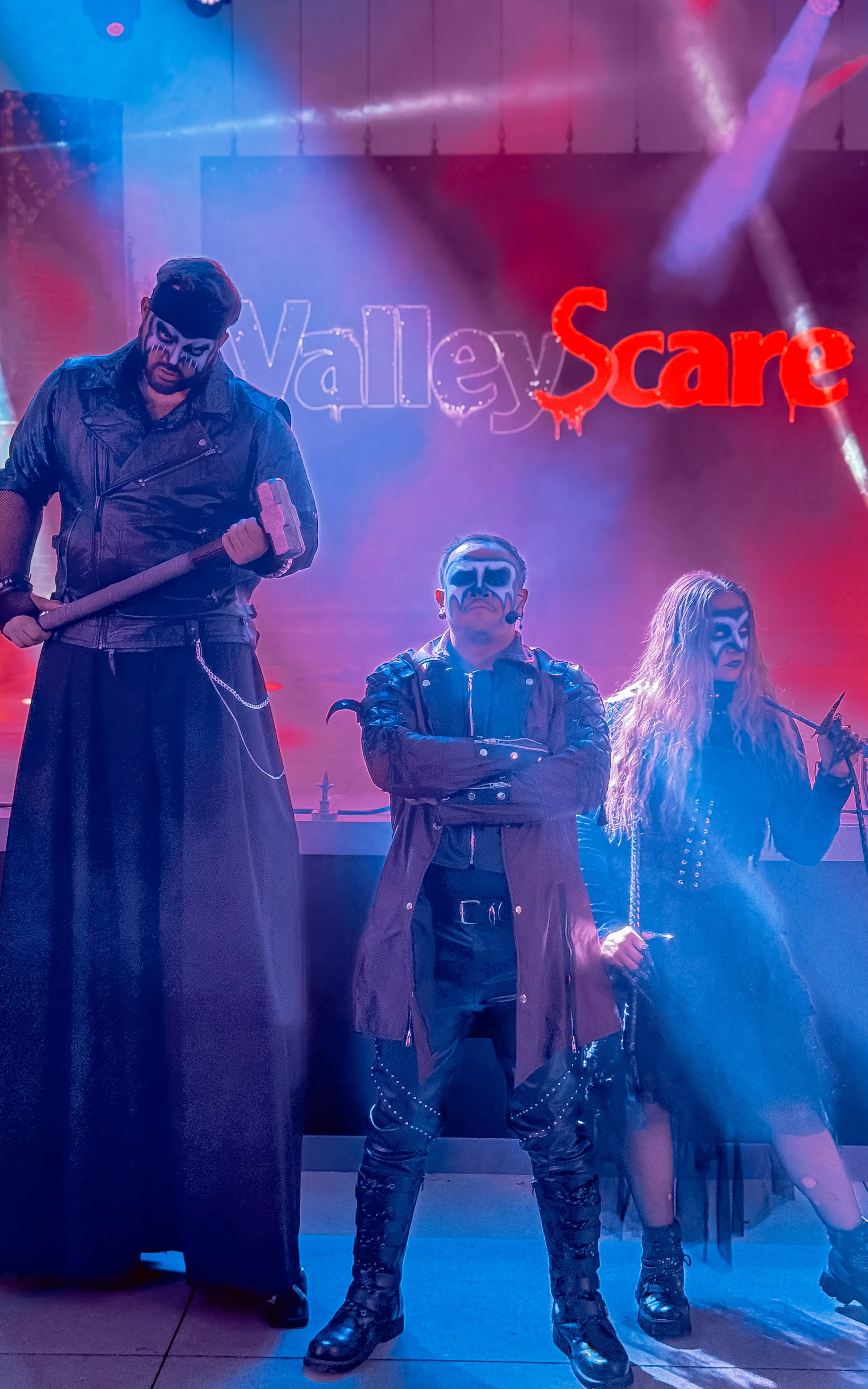 Three people dressed in dark, gothic costumes with face paint standing in front of a neon sign that says "Valley Scare". The person on the left is holding a hammer, and the two on the right are holding knives. The scene is lit with purple and red lig