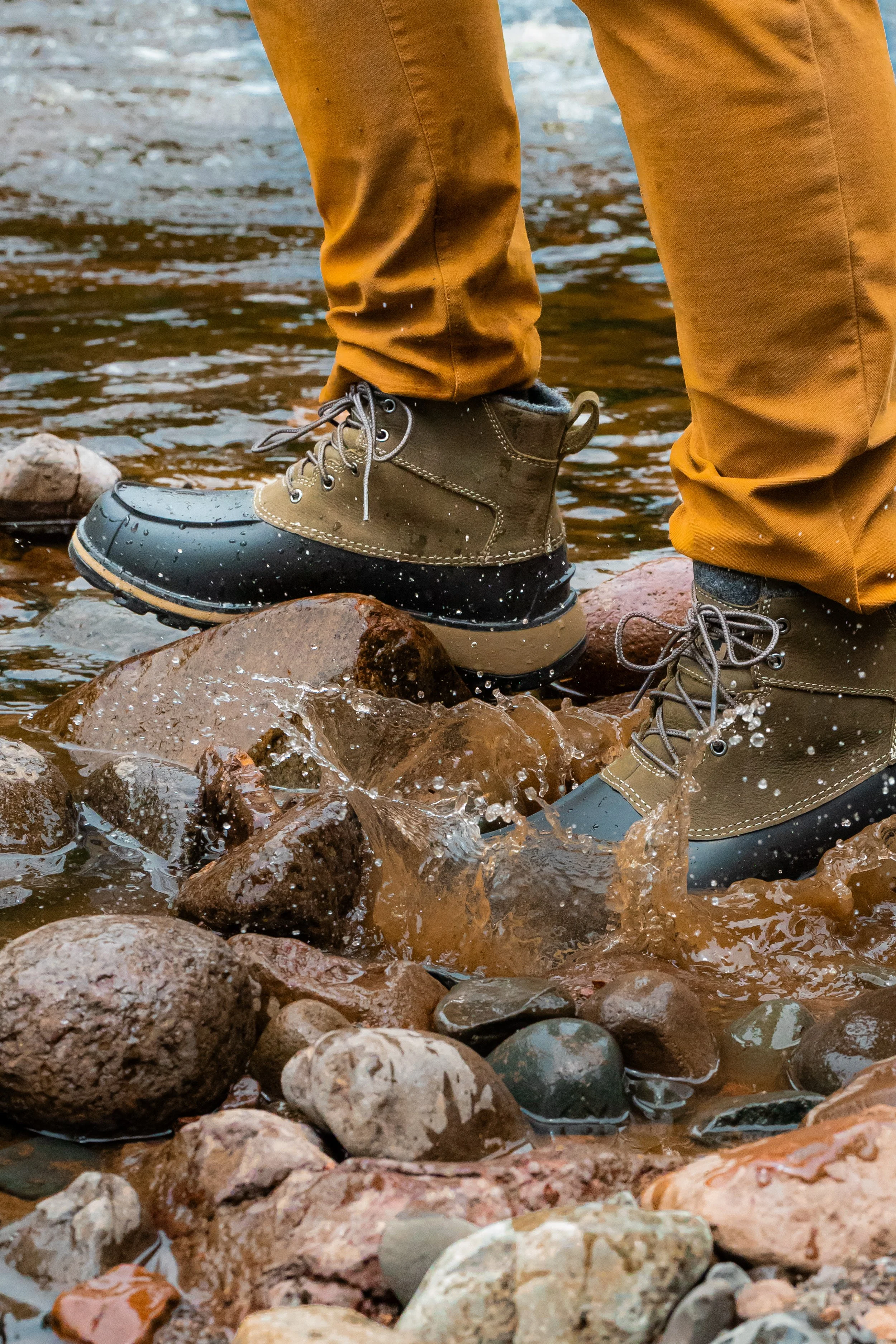 Person wearing waterproof boots and orange pants standing in shallow water over rocks.