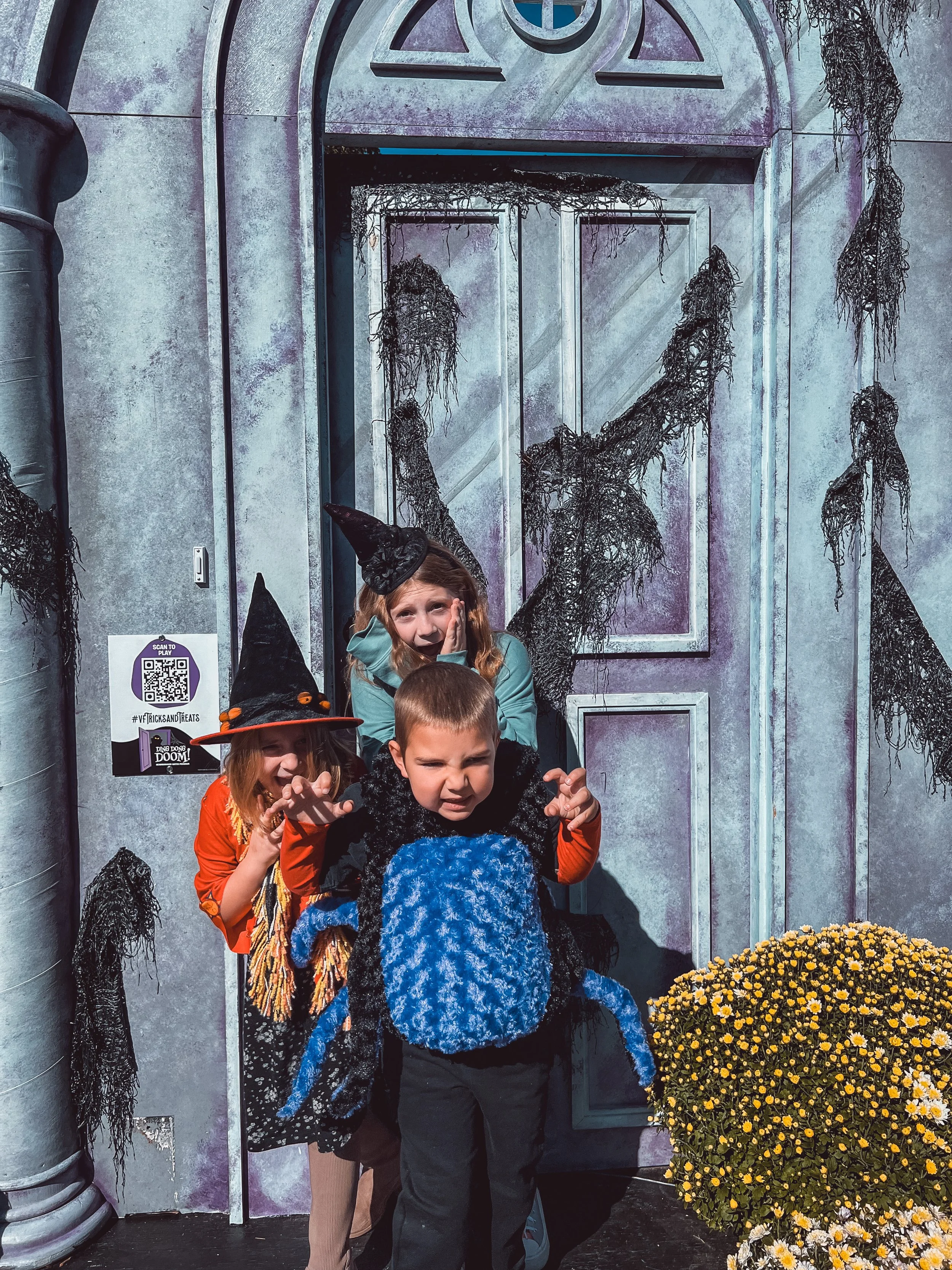 Three children dressed in Halloween costumes standing outside a spooky, painted door with black webs and a large pumpkin decoration nearby.