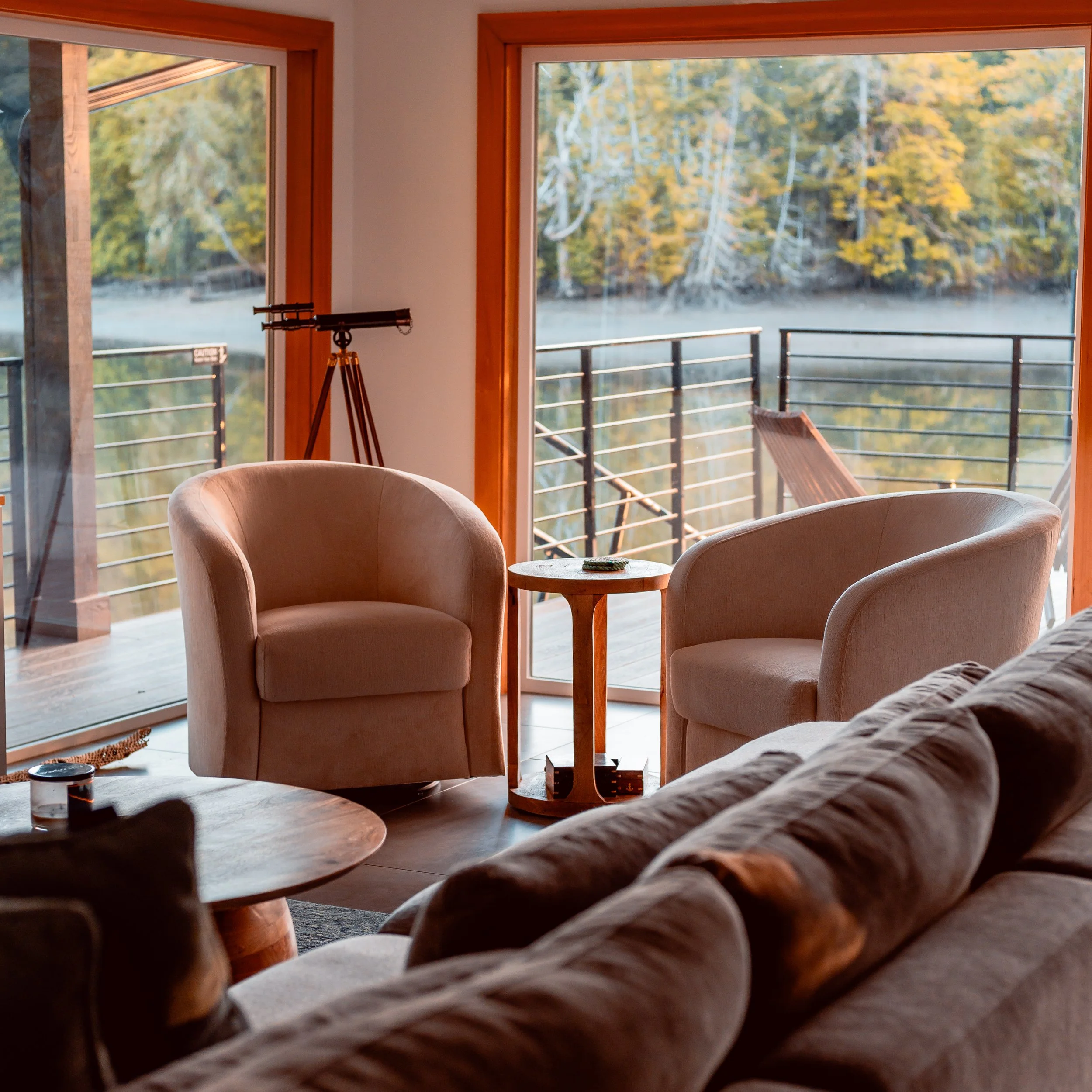 Living room with beige armchairs, a wooden side table, and large glass sliding doors leading to a balcony overlooking a lake and trees with fall foliage.